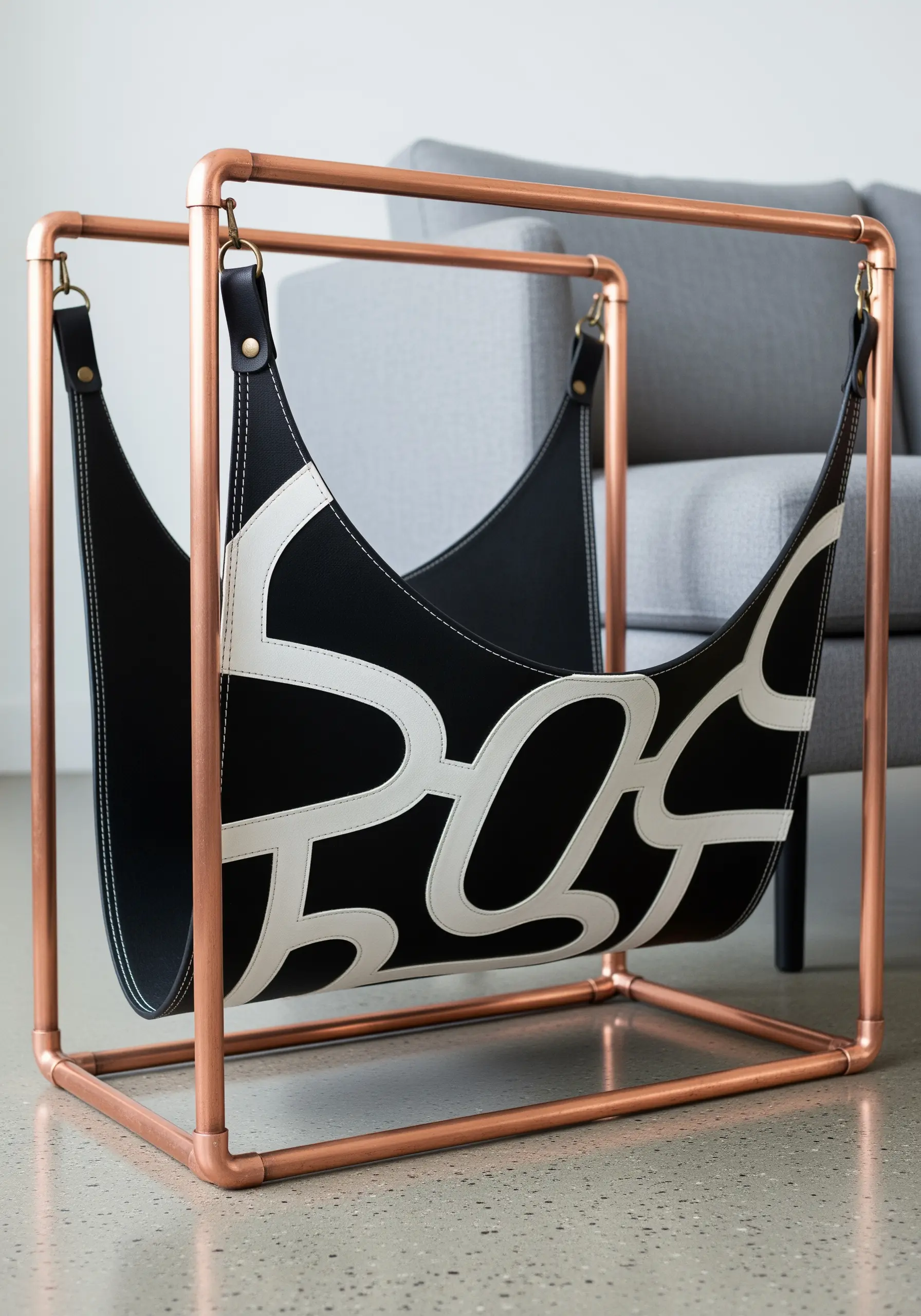 A modern magazine rack with a copper pipe frame and a black leather sling with white appliqué.