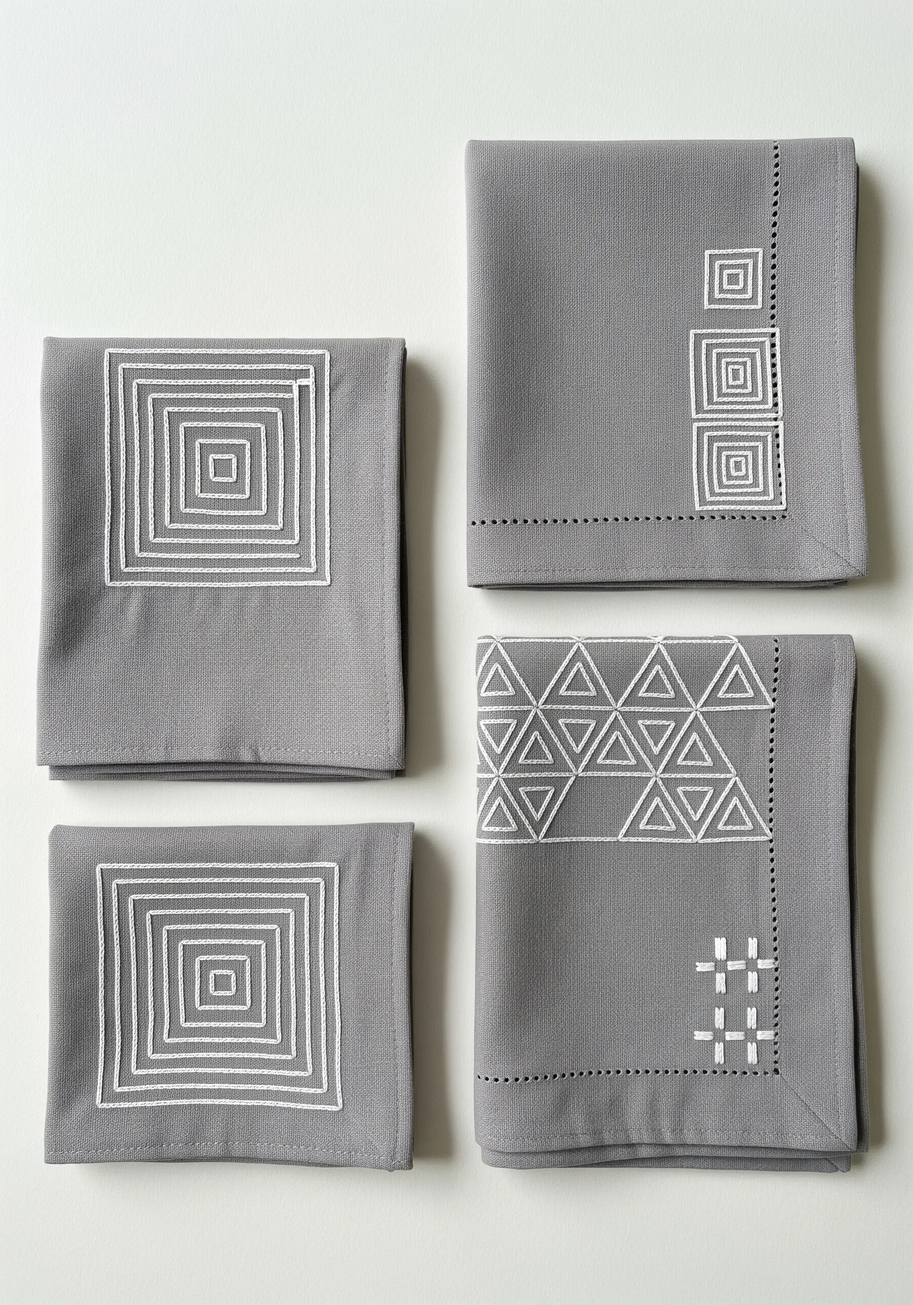 Four grey napkins with different geometric patterns embroidered in white thread.