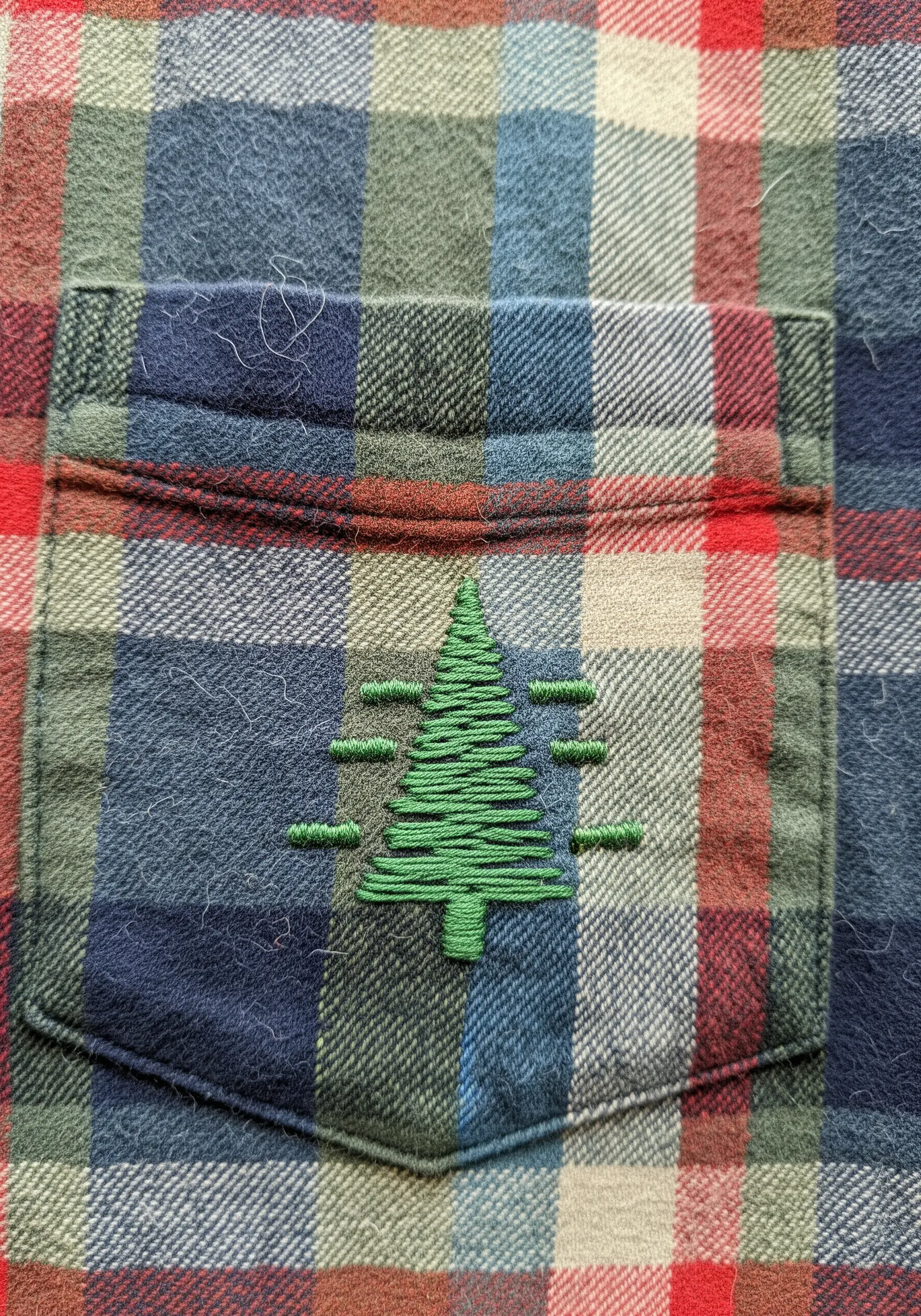 A green embroidered pine tree on the pocket of a red, green, and blue plaid flannel shirt.