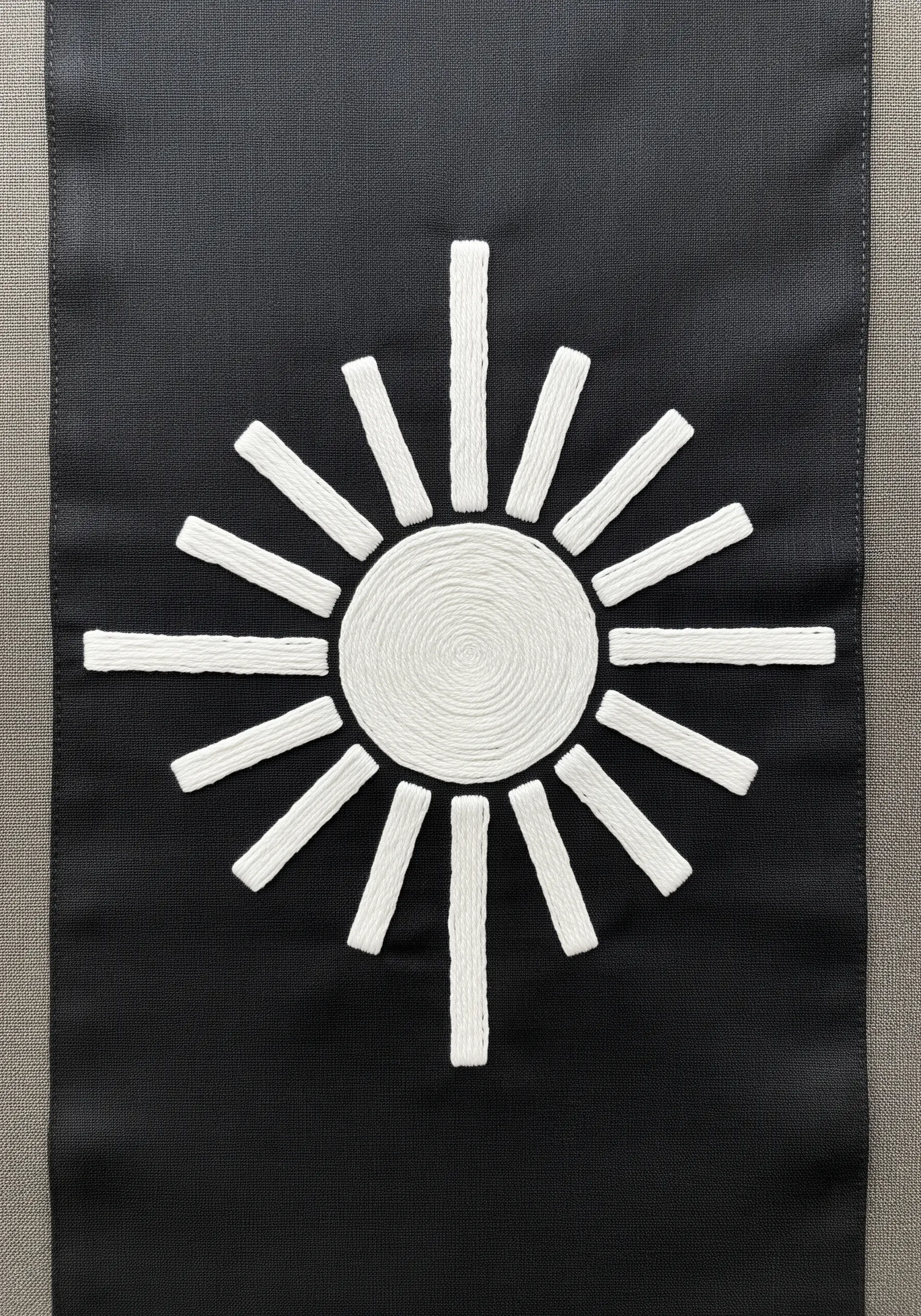 A bold sun motif made from couched white rope on a black fabric table runner.