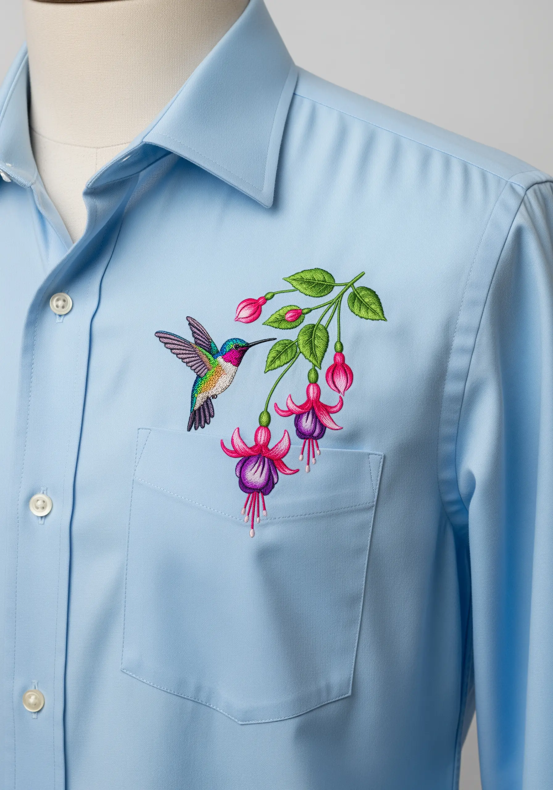 A colorful hummingbird hovering by fuchsia flowers, embroidered on a light blue shirt.
