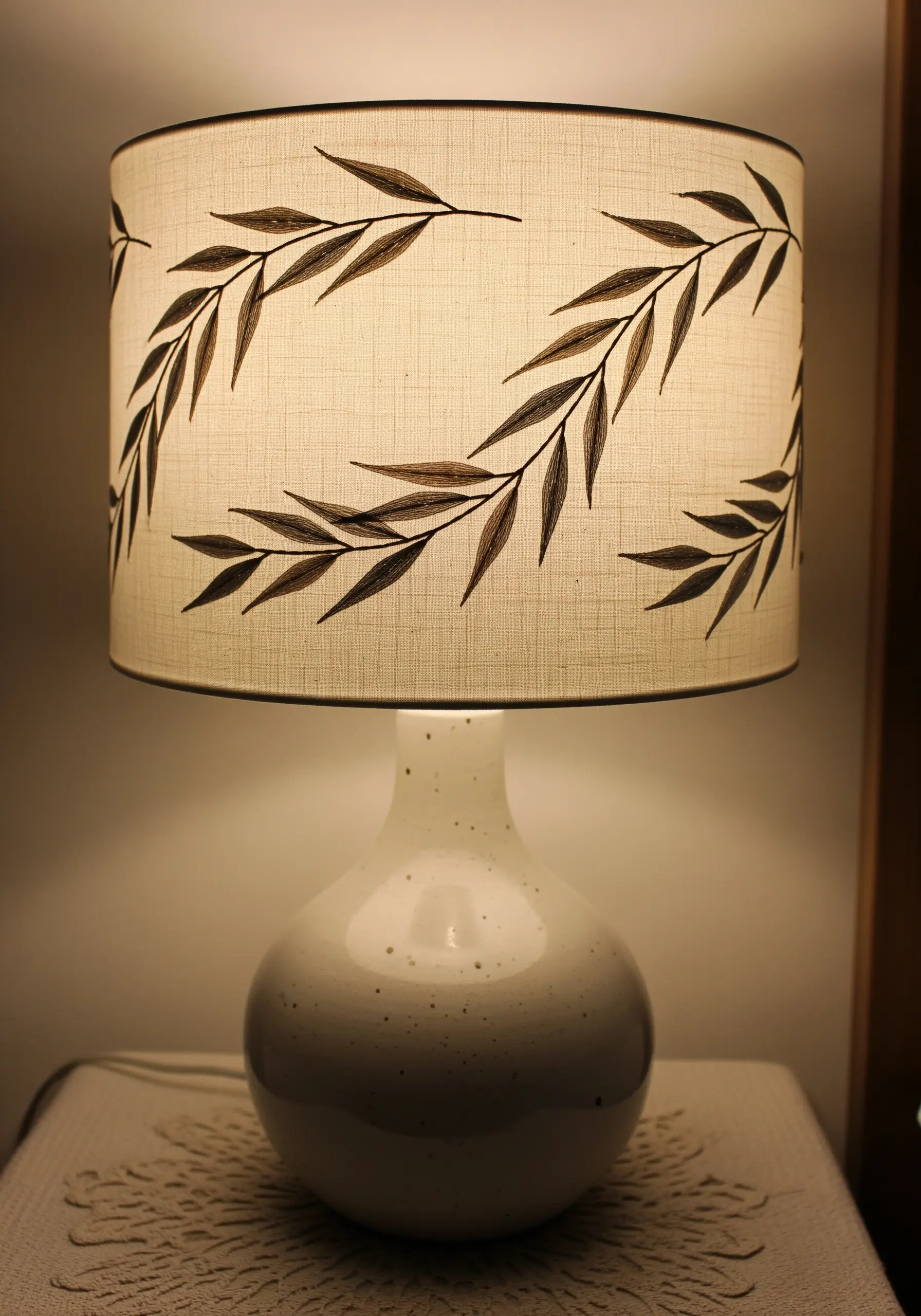 A cream-colored lampshade with a dark brown embroidered willow branch pattern, lit from within.