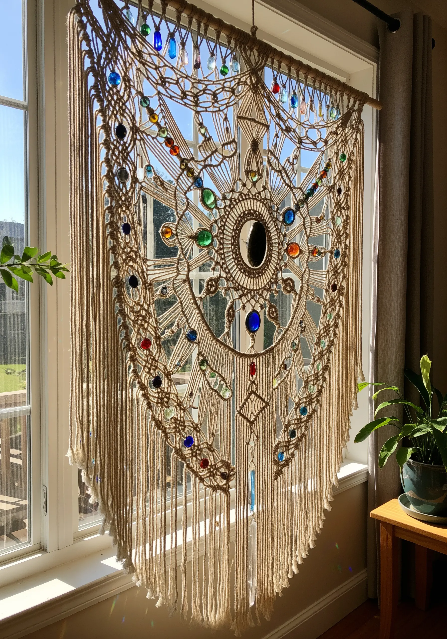 A detailed macrame wall hanging in a window, embedded with colorful glass beads and a central mirror.