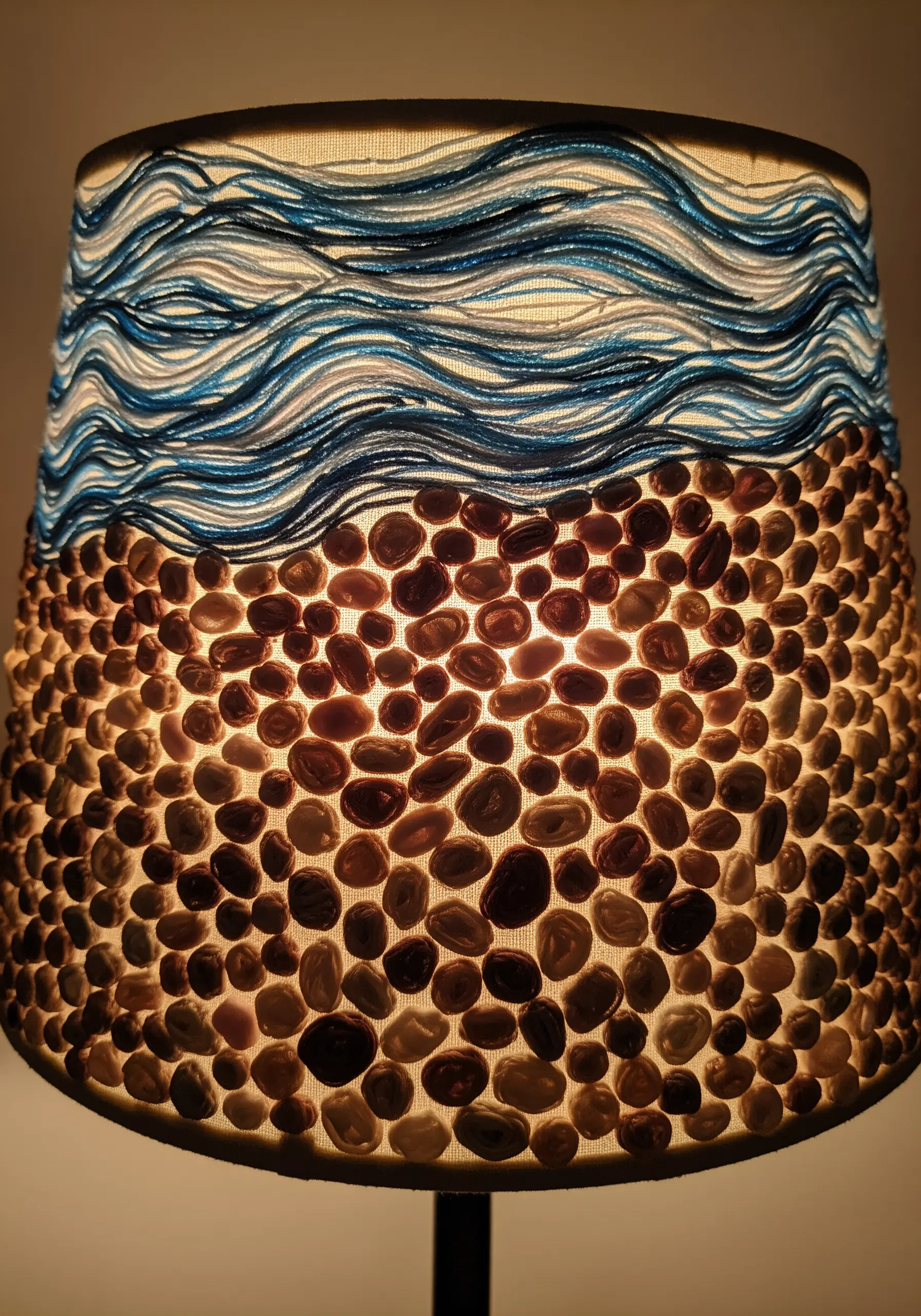 A lit lampshade decorated with embroidered blue waves on top and a mosaic of polished stones below.