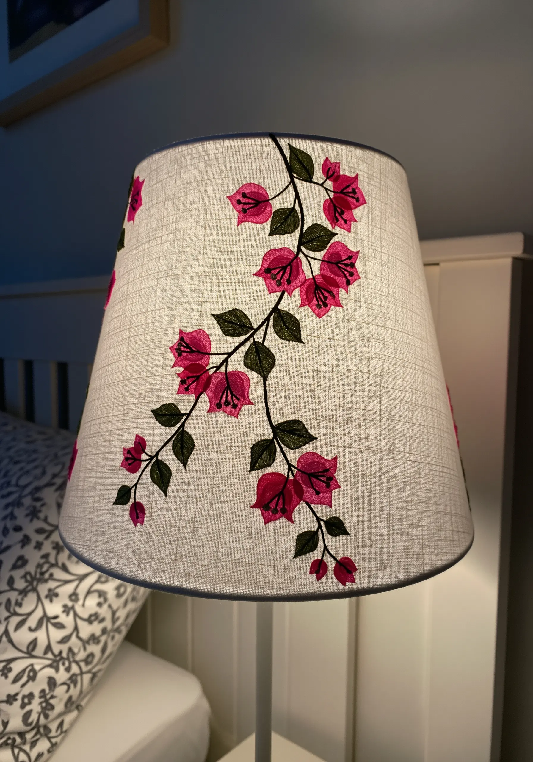 A fabric lampshade illuminated from within, showcasing an embroidered pattern of pink bougainvillea.