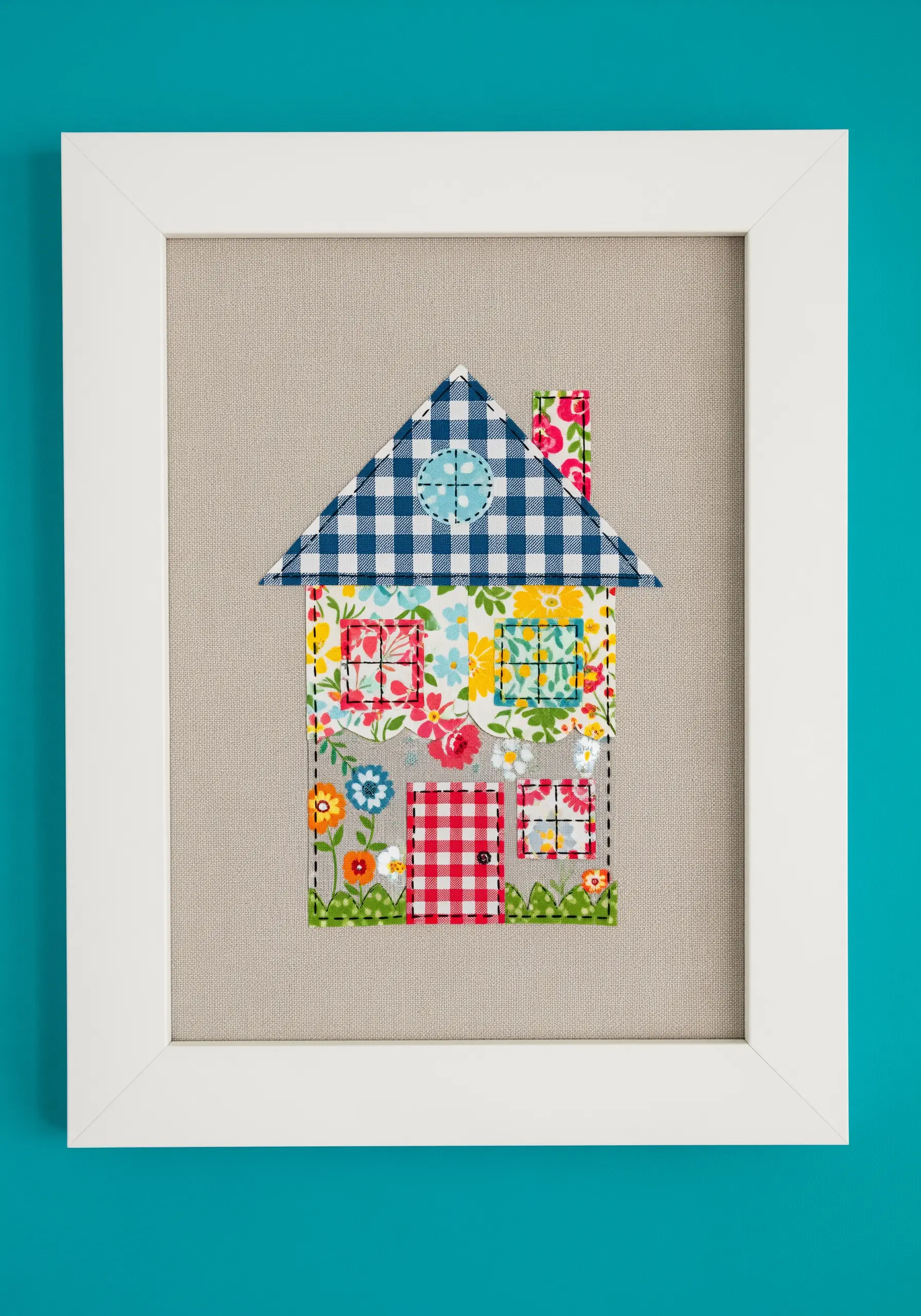 A framed art piece showing a house made from various patterned fabric scraps.