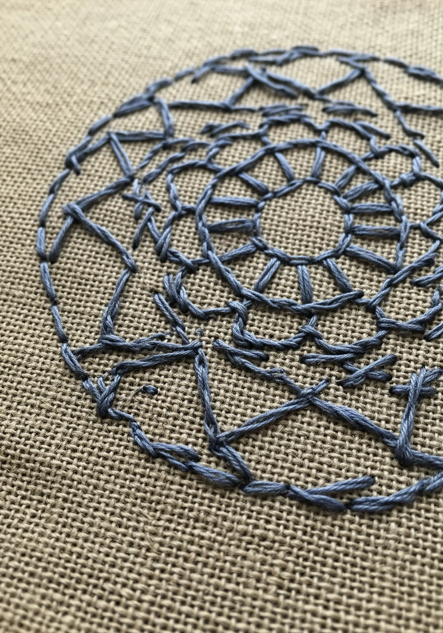 A delicate, open-weave mandala stitched on coarse, natural burlap fabric.