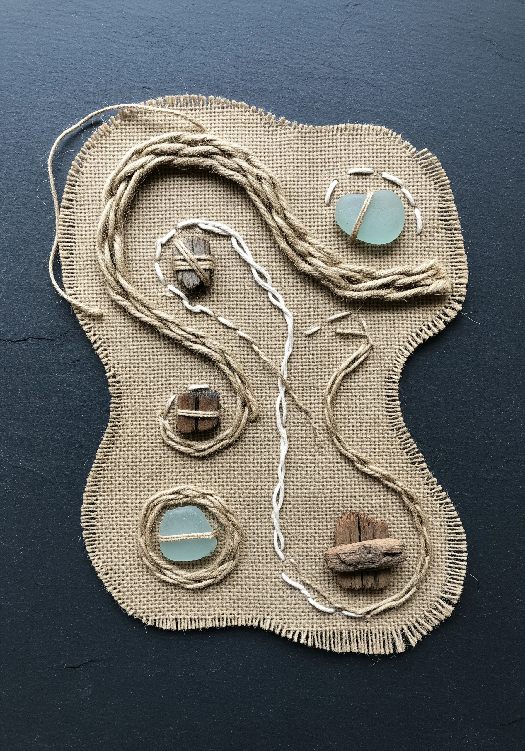 A mixed-media piece on burlap featuring sea glass and driftwood secured with couched twine.