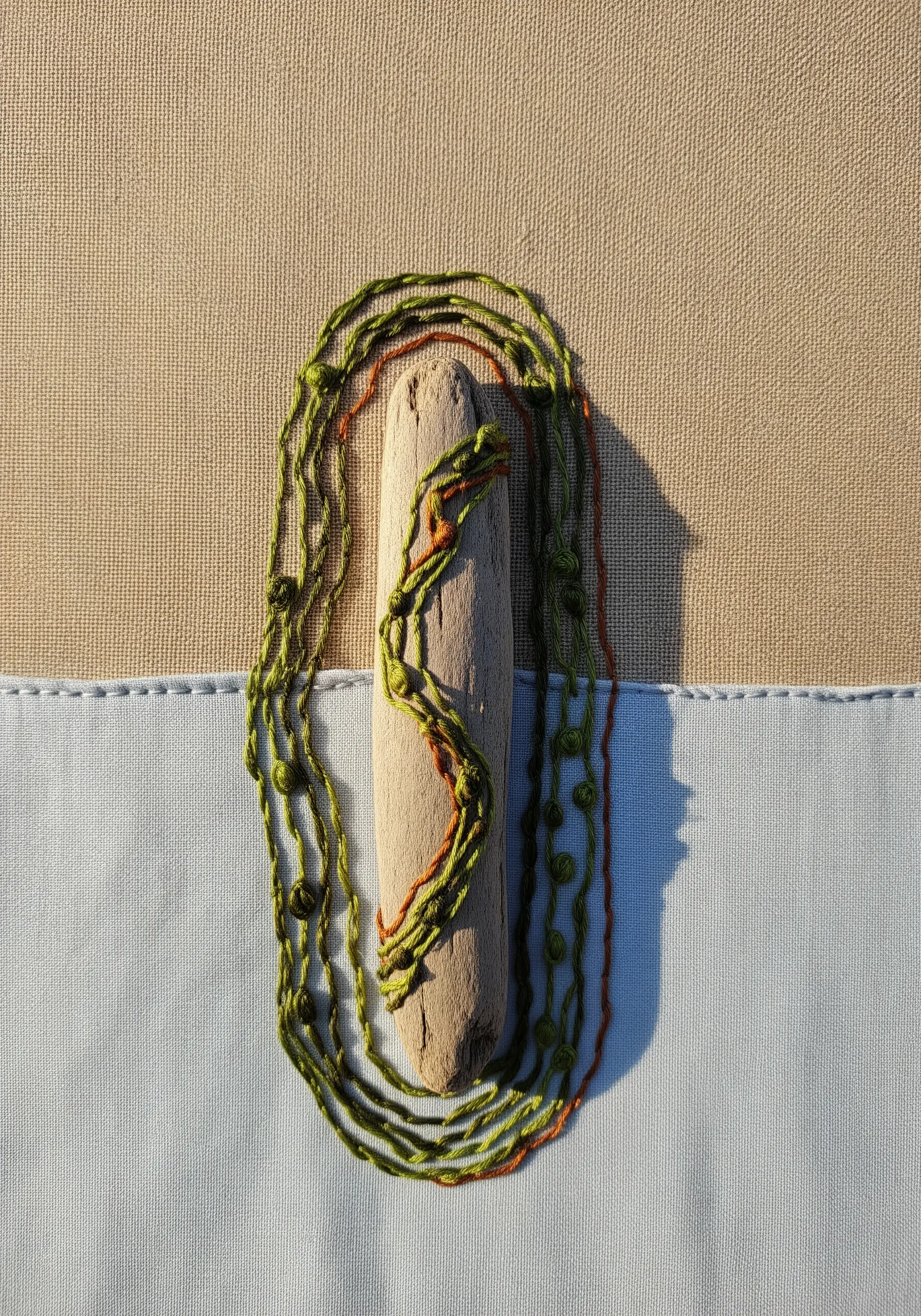 A piece of driftwood secured to canvas and integrated with wrapped green and orange threads.