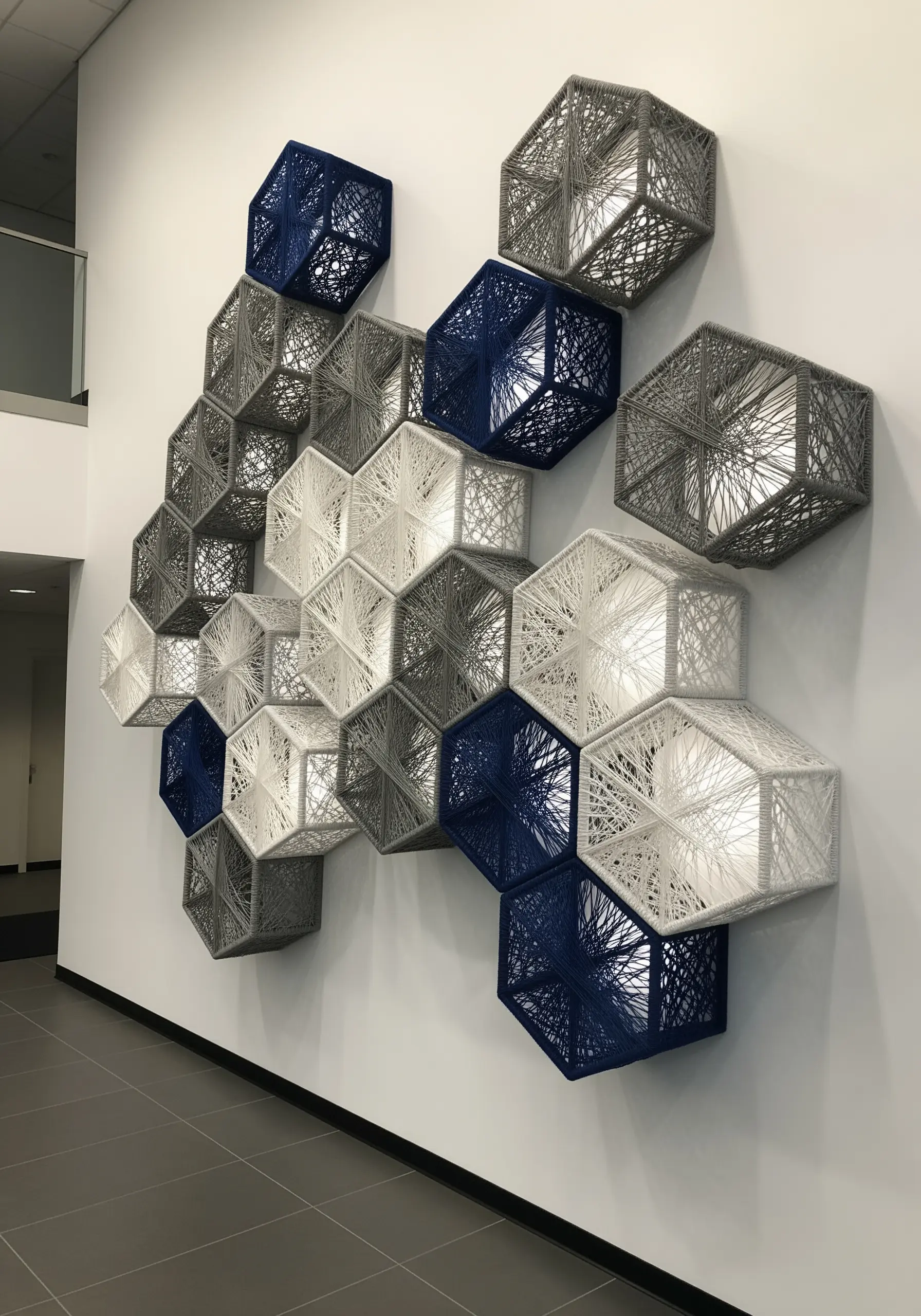 A wall installation of 3D hexagonal frames connected by webs of white, grey, and blue string.