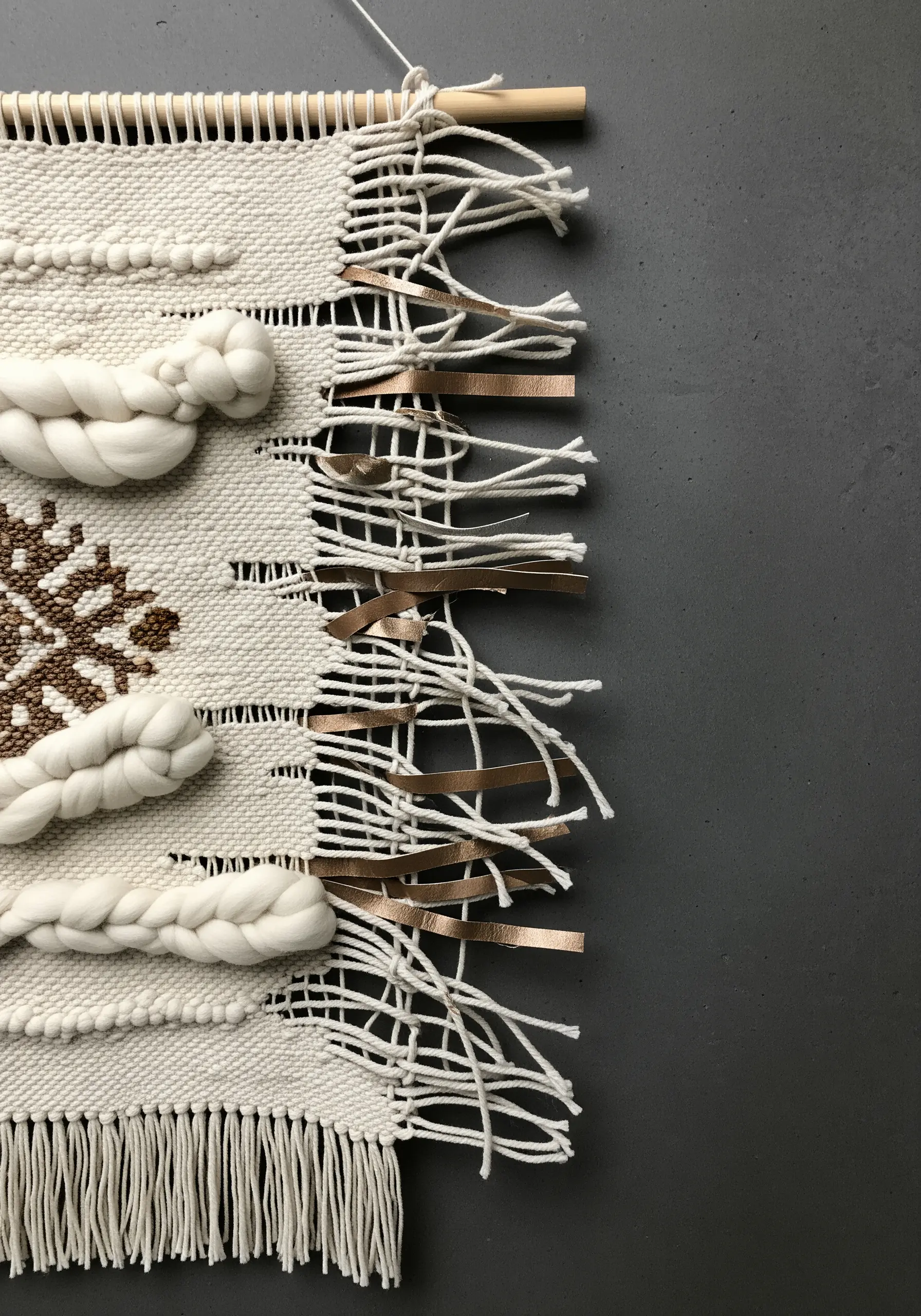A small weaving combining chunky white wool, brown yarn, and metallic leather strips.