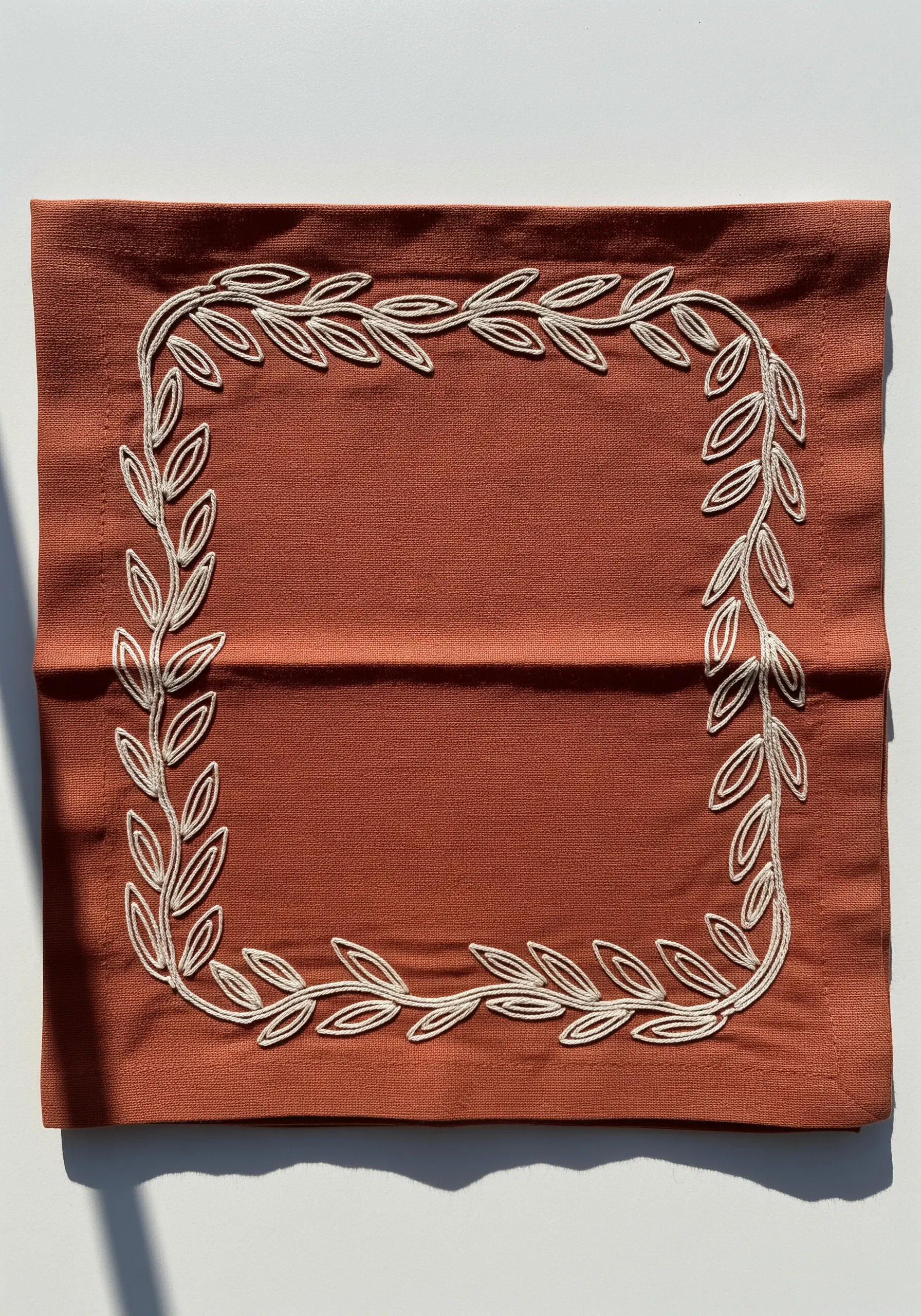 A full border of laurel leaves created with a raised, couched white cord on a rust napkin.