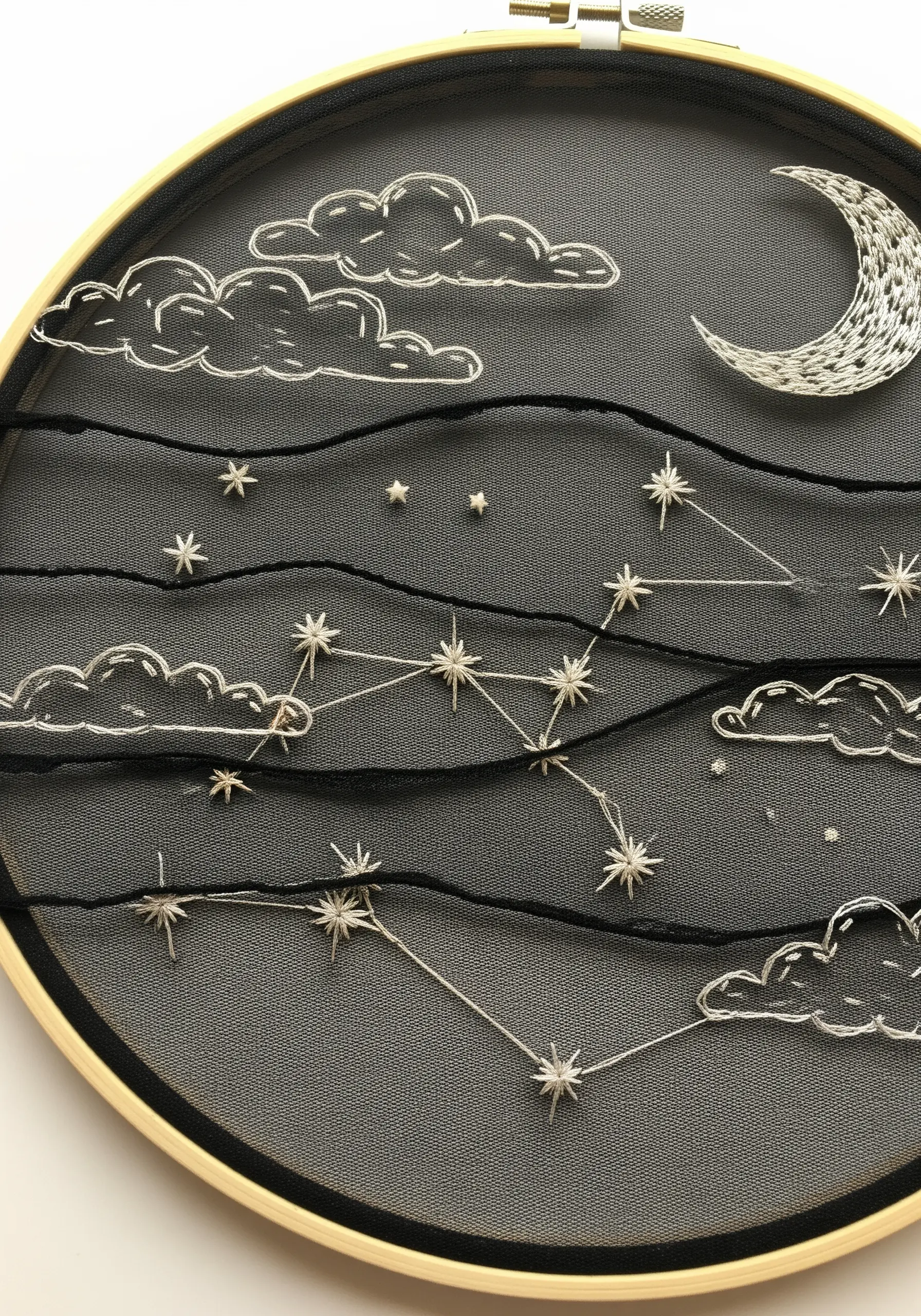 A night sky with clouds, moon, and constellations embroidered on black tulle within a hoop.