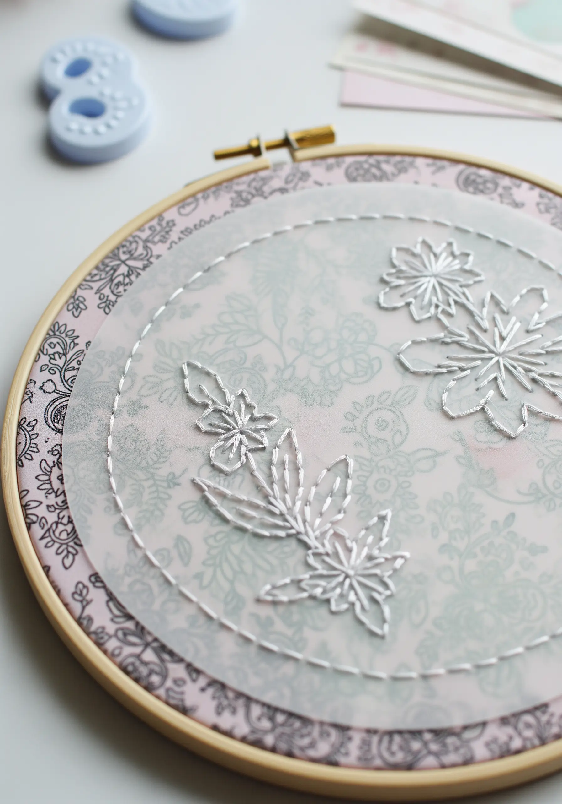 White floral embroidery on a sheer fabric, layered over a soft pink patterned paper in a hoop.