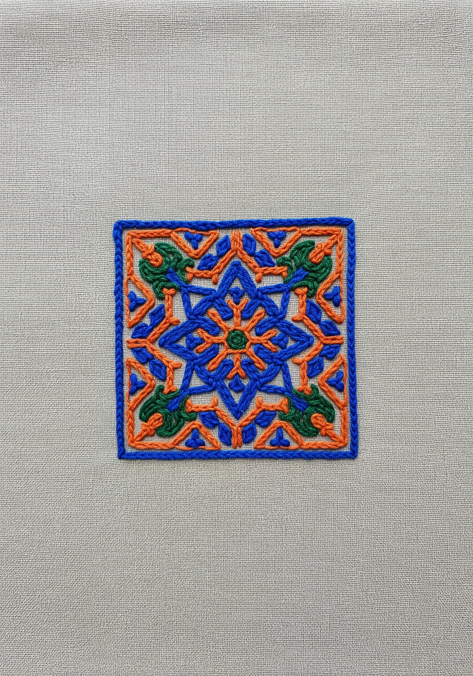 A single, intricate embroidered tile in blue, orange, and green on linen fabric.