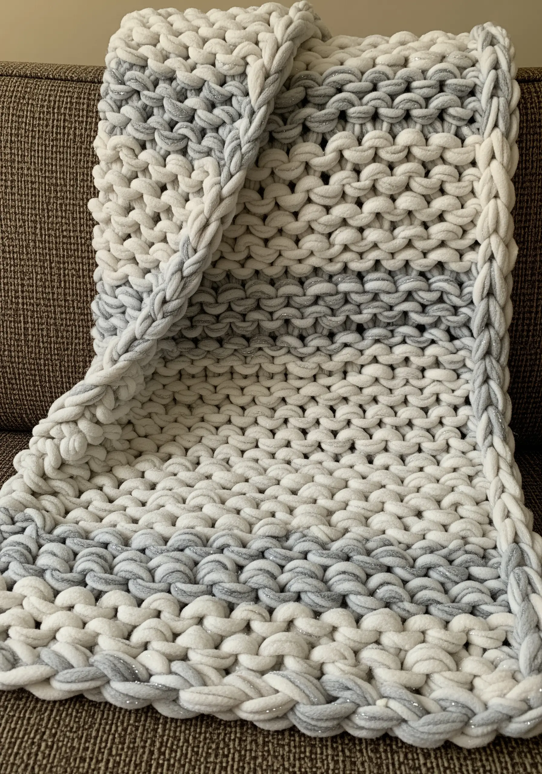 A chunky arm-knitted blanket in grey and white chenille yarn on a sofa.