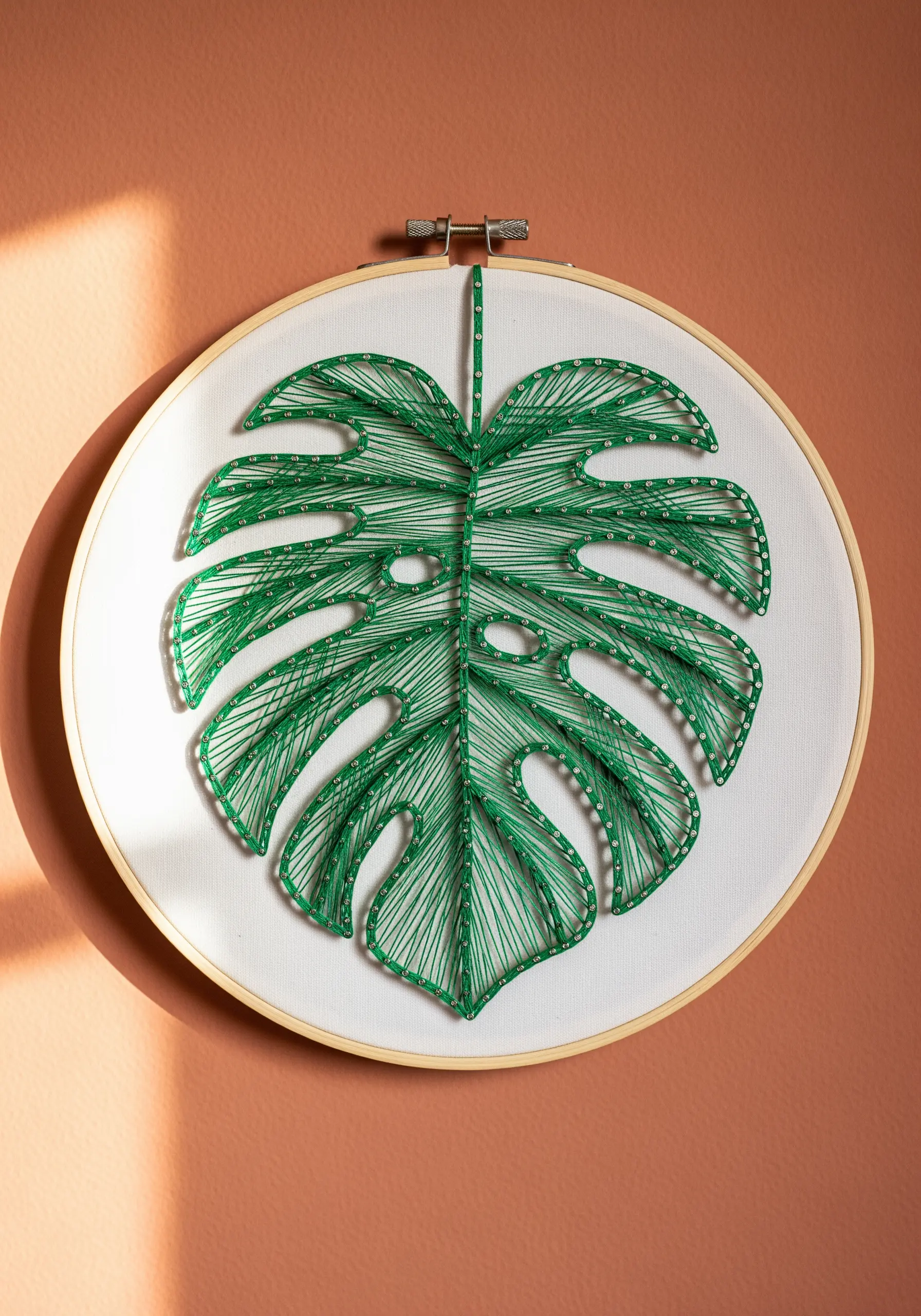 A string art monstera leaf in a hoop, with green thread stretched between anchor points.