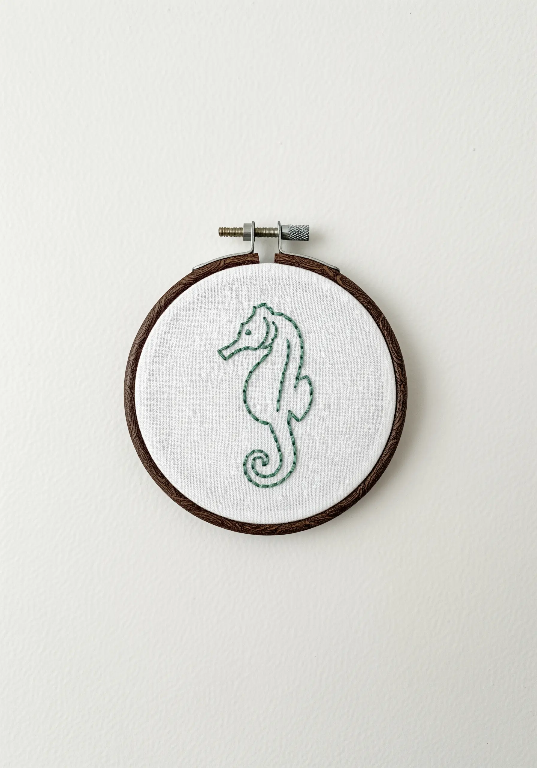 Minimalist seahorse outline embroidered in green thread on white fabric in a small hoop.