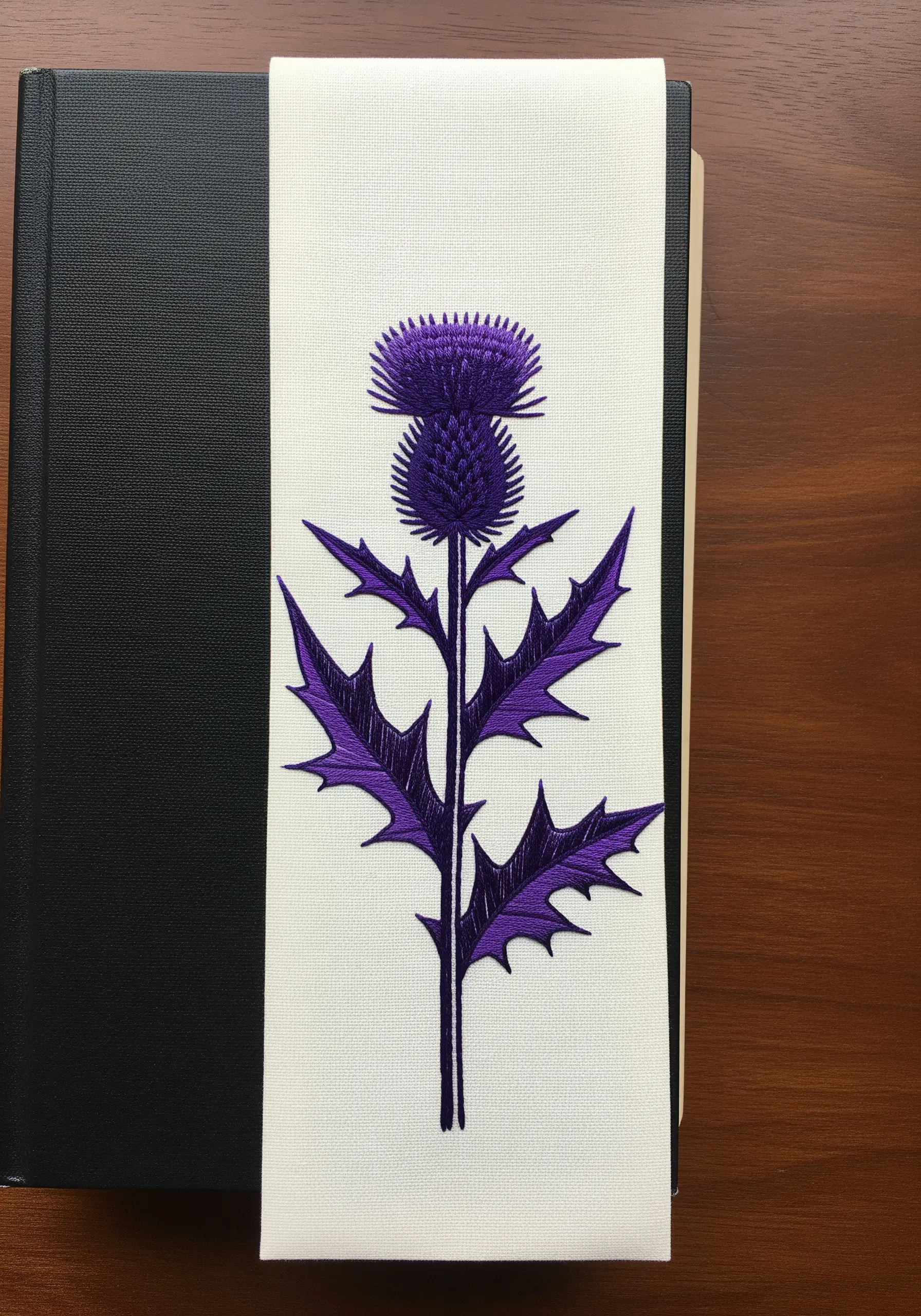 A bold purple thistle with sharp leaves, embroidered with satin stitch on a white bookmark.