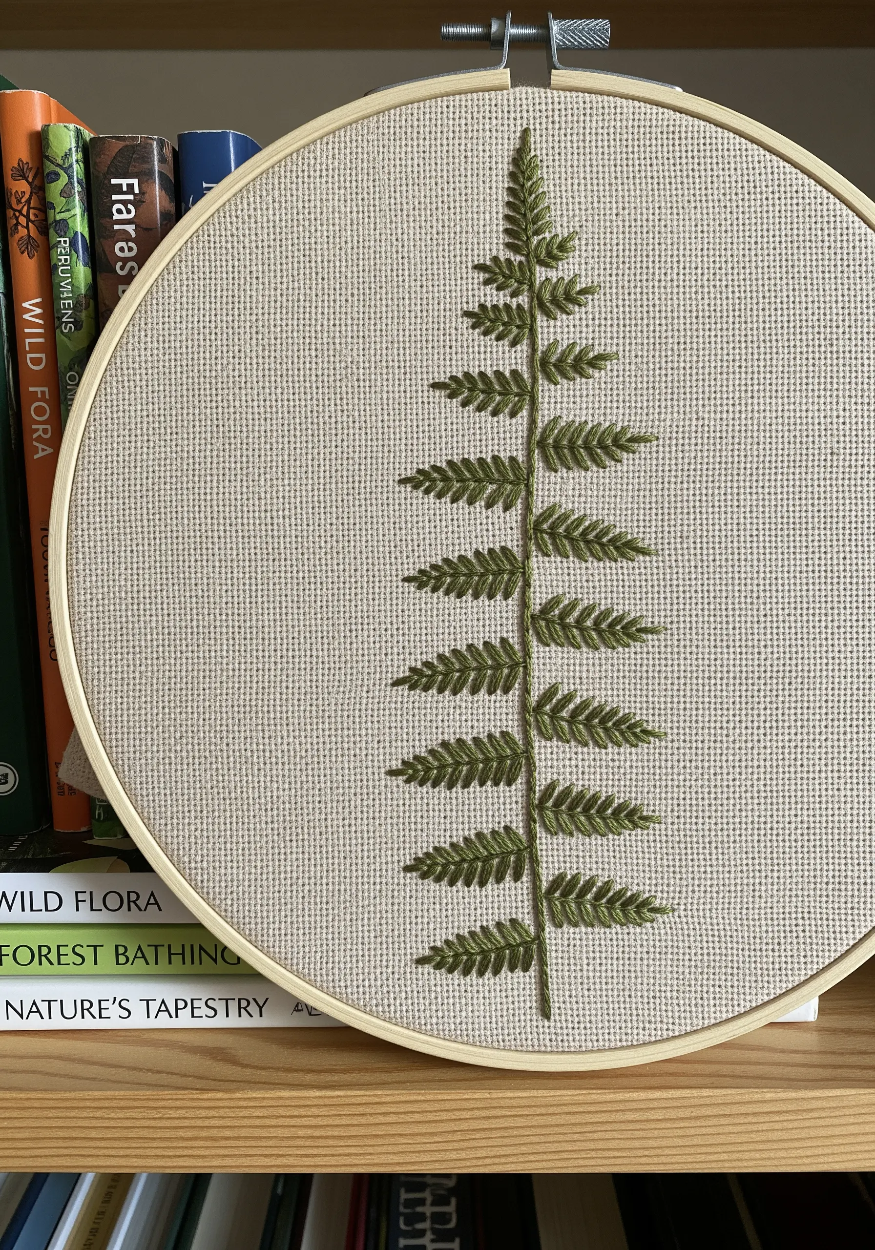 A single, symmetrical embroidered fern frond in olive green on oatmeal-colored Aida fabric.