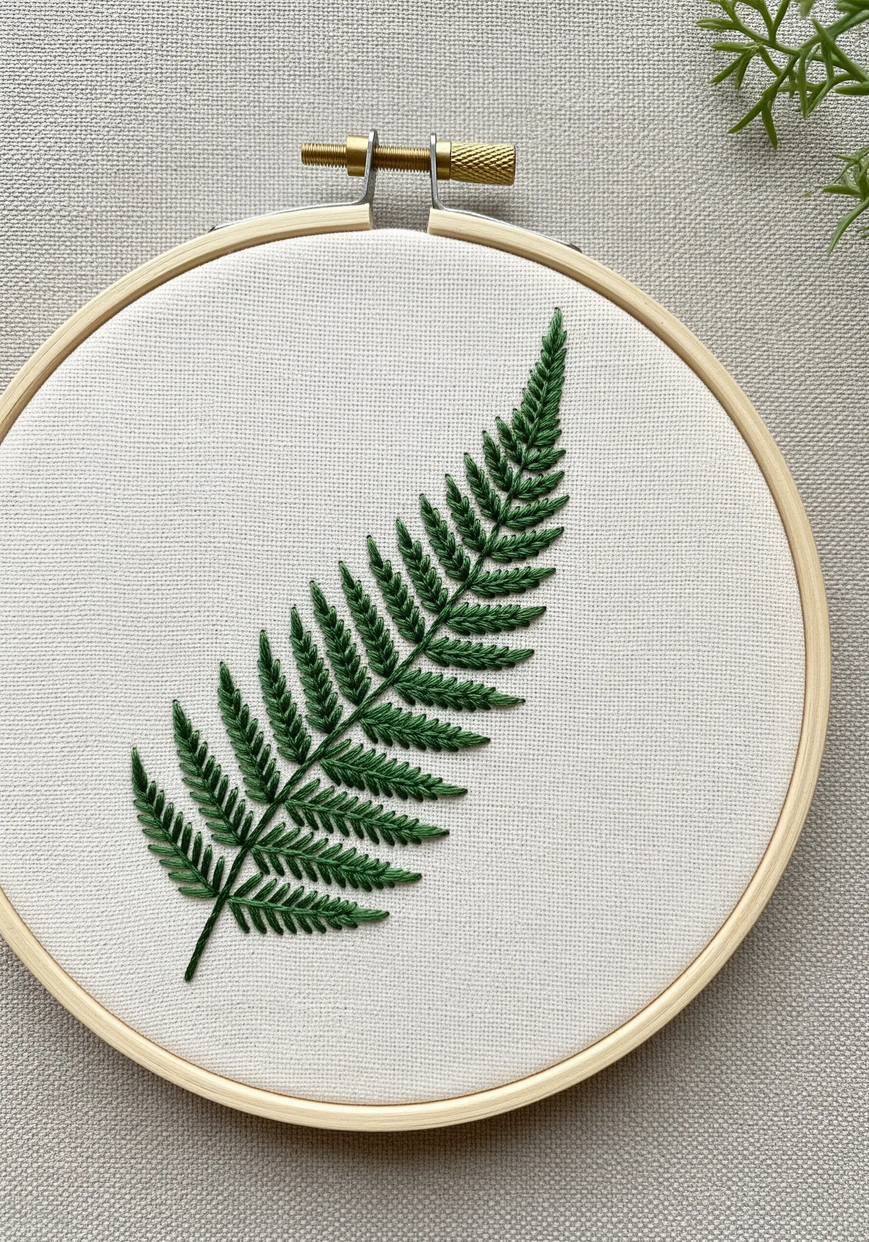 A perfectly stitched fern leaf in dark green thread on off-white fabric, showing precise stitches.