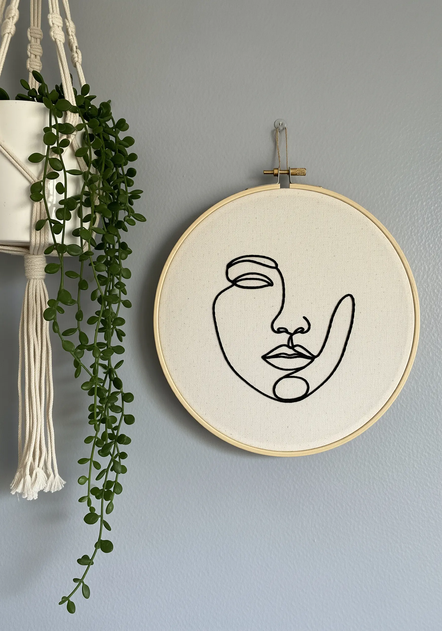 Minimalist line art face embroidered in thick black thread on a neutral canvas hoop.