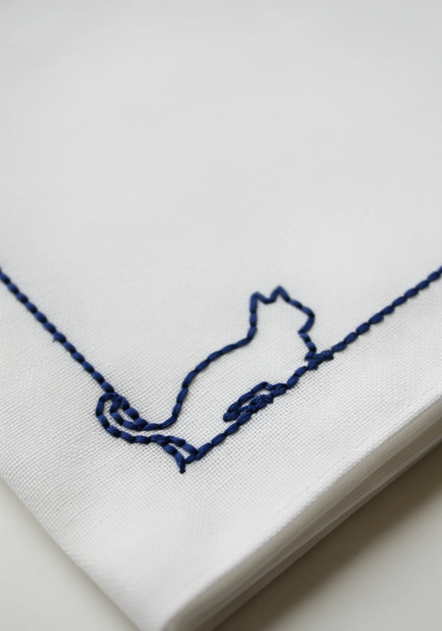 The outline of a sitting cat stitched in a continuous line of dark blue thread on white fabric.
