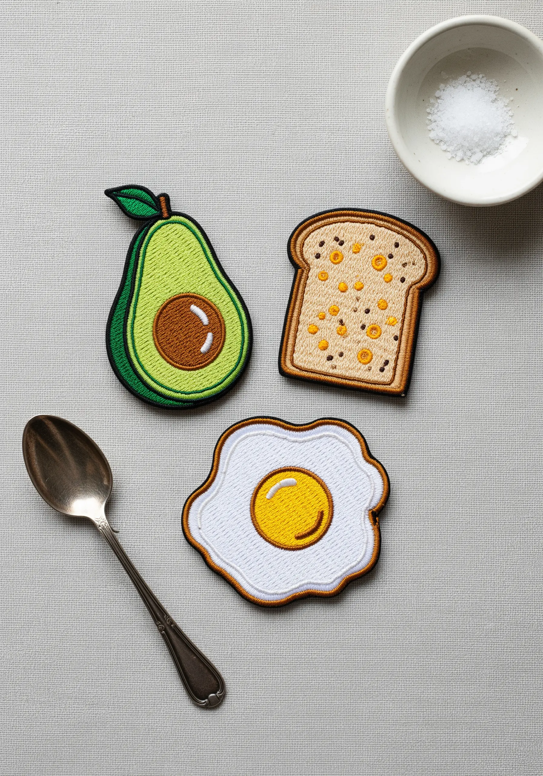 Three embroidered patches shaped like an avocado, toast, and a fried egg on a neutral surface.