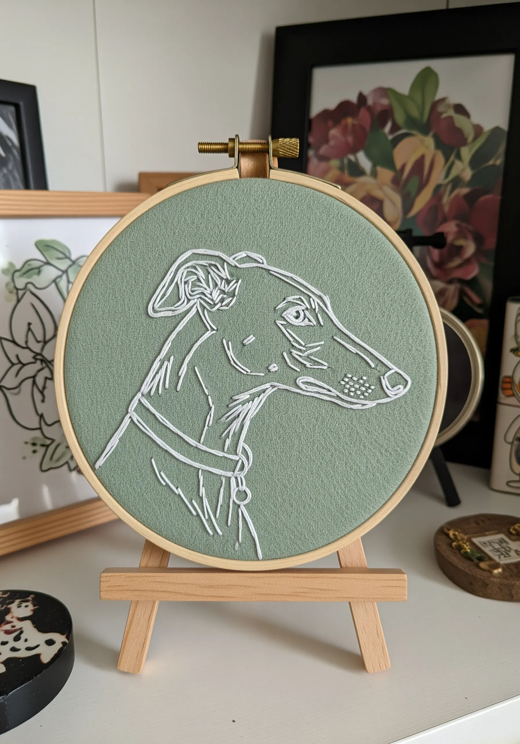 White thread line-art portrait of a greyhound on a sage green background.