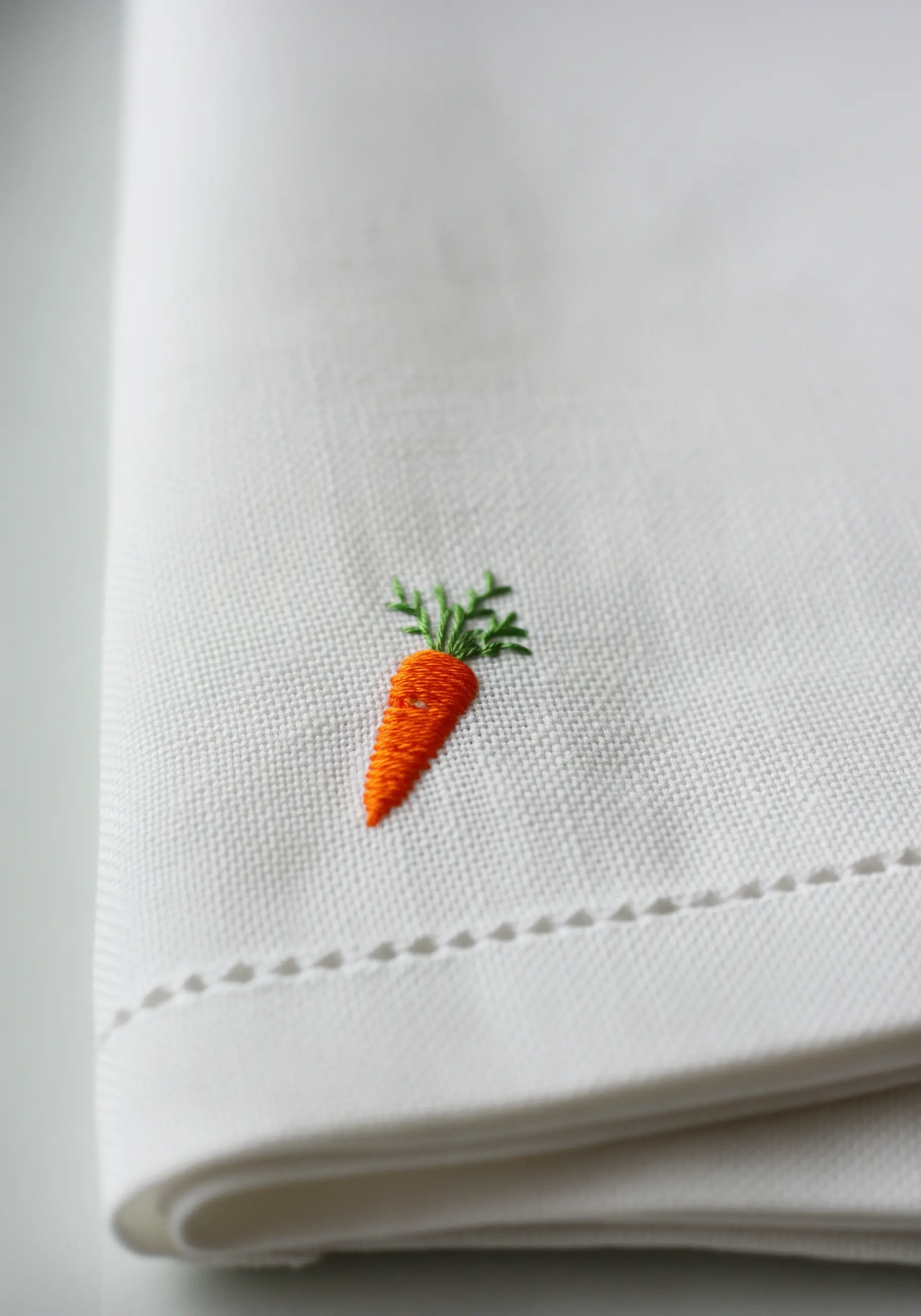 A tiny embroidered carrot with satin stitch body and detached chain stitch leaves on a white linen napkin.
