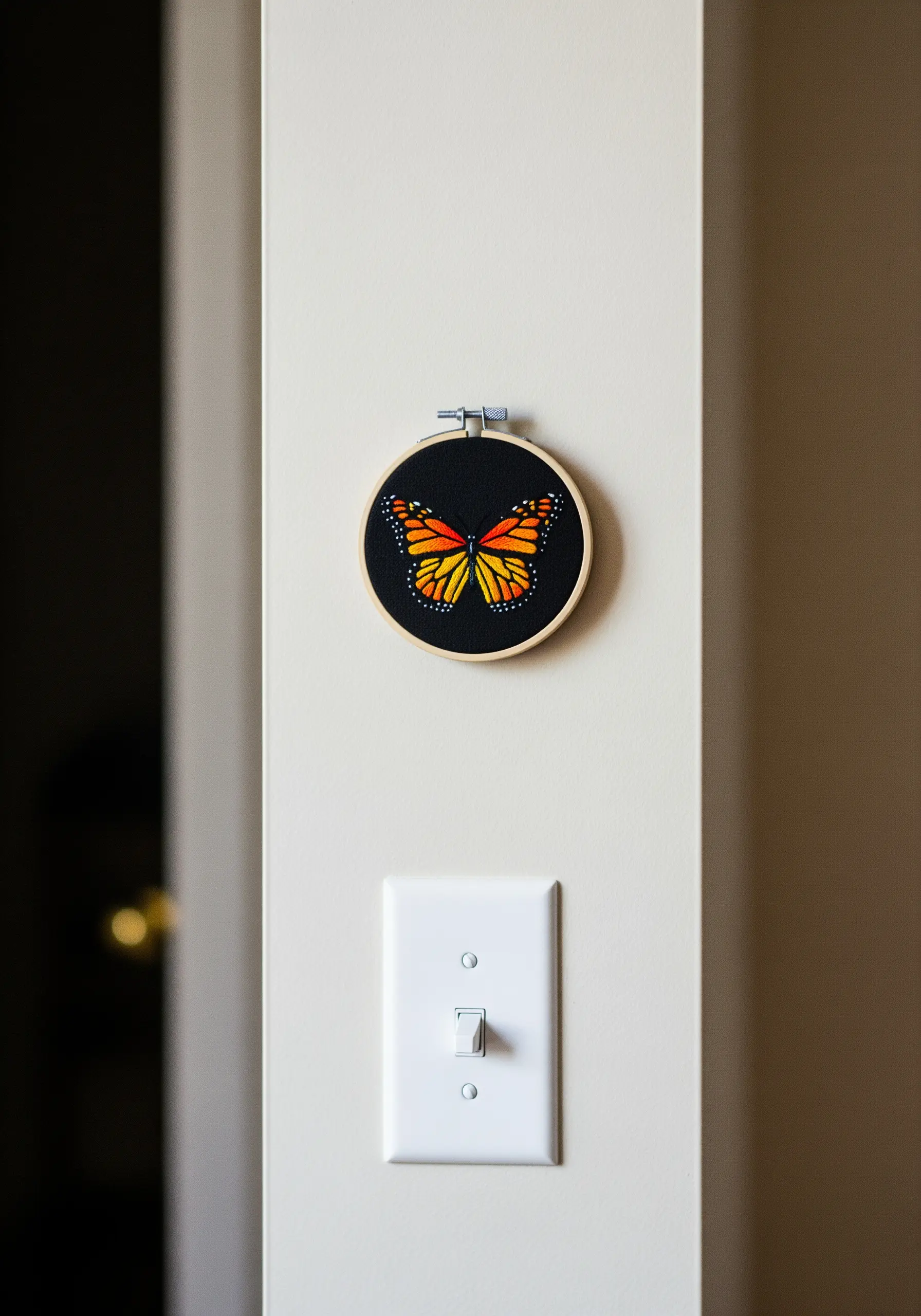 A small embroidery hoop with a vibrant monarch butterfly stitched on black fabric, near a light switch.