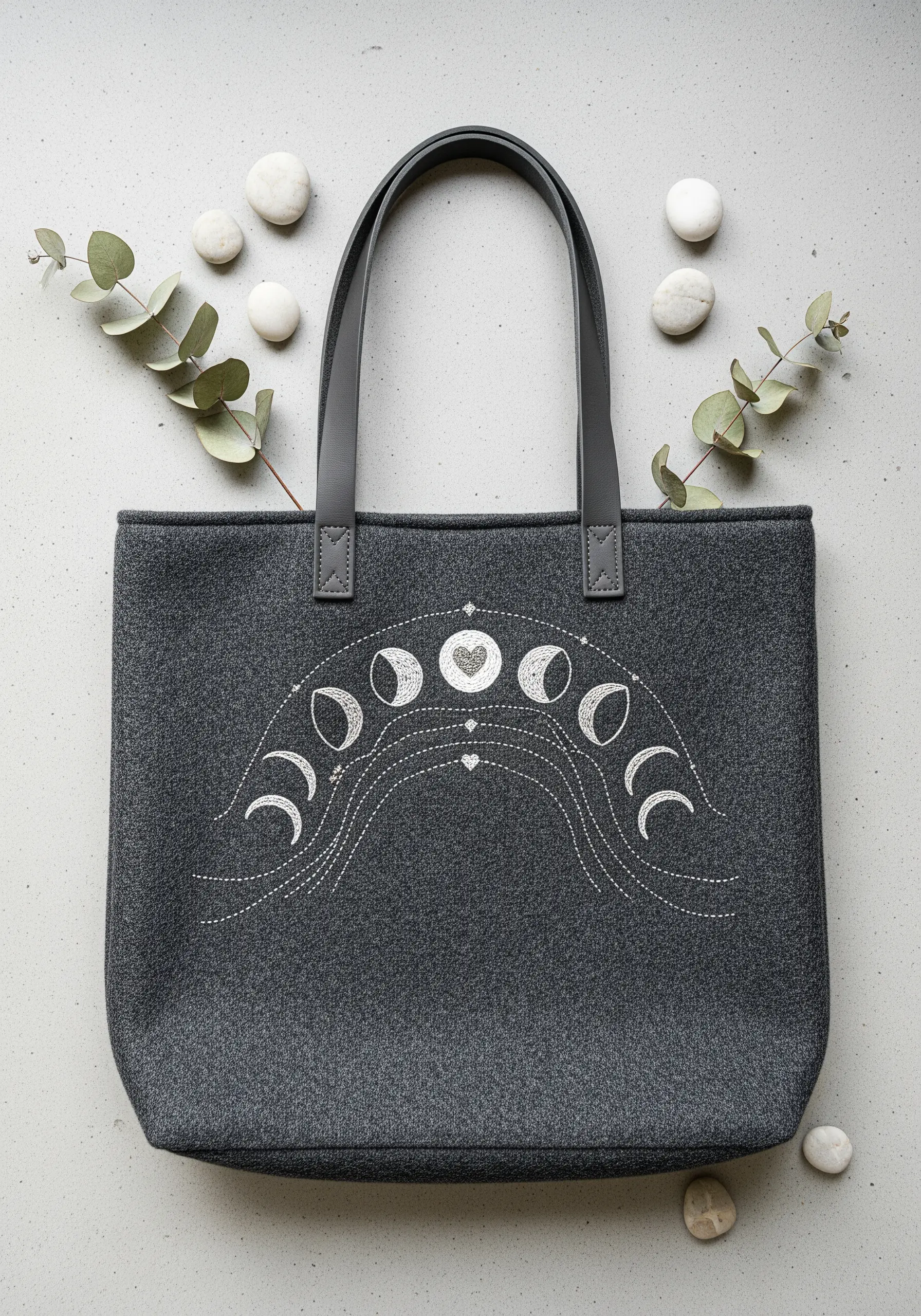 A minimalist arc of moon phases embroidered in white thread on a dark gray felt tote bag.