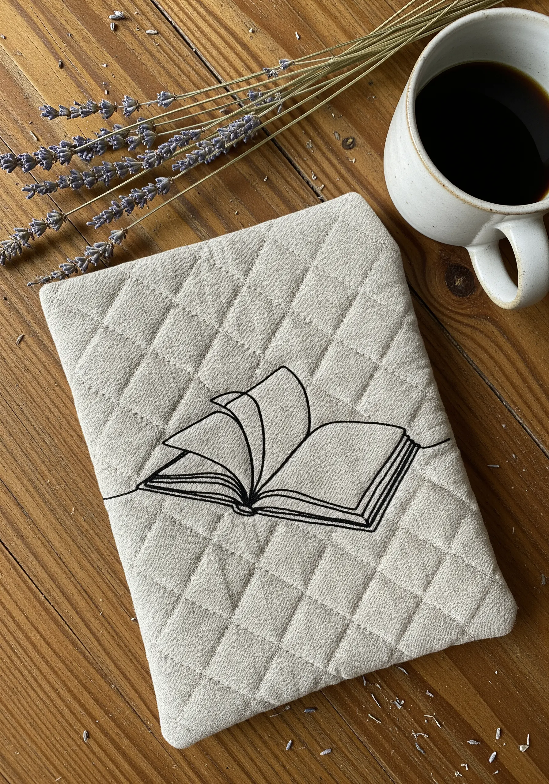 Black line art embroidery of an open book on beige quilted fabric sleeve