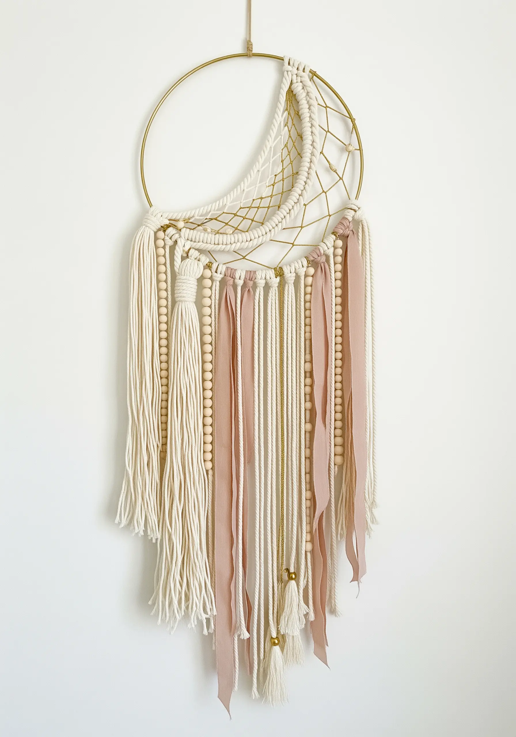 A modern macrame dreamcatcher on a crescent moon frame with ribbons and wooden beads.