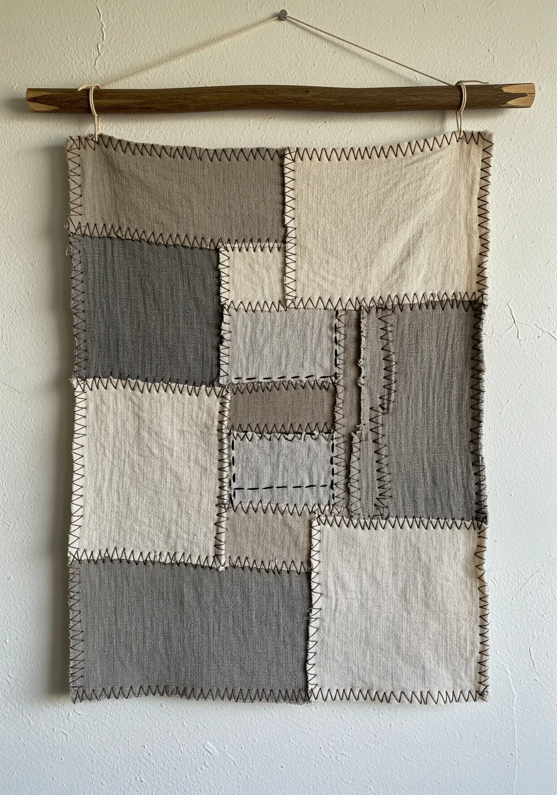 A patchwork wall hanging of neutral fabric squares joined with a prominent dark zigzag stitch.