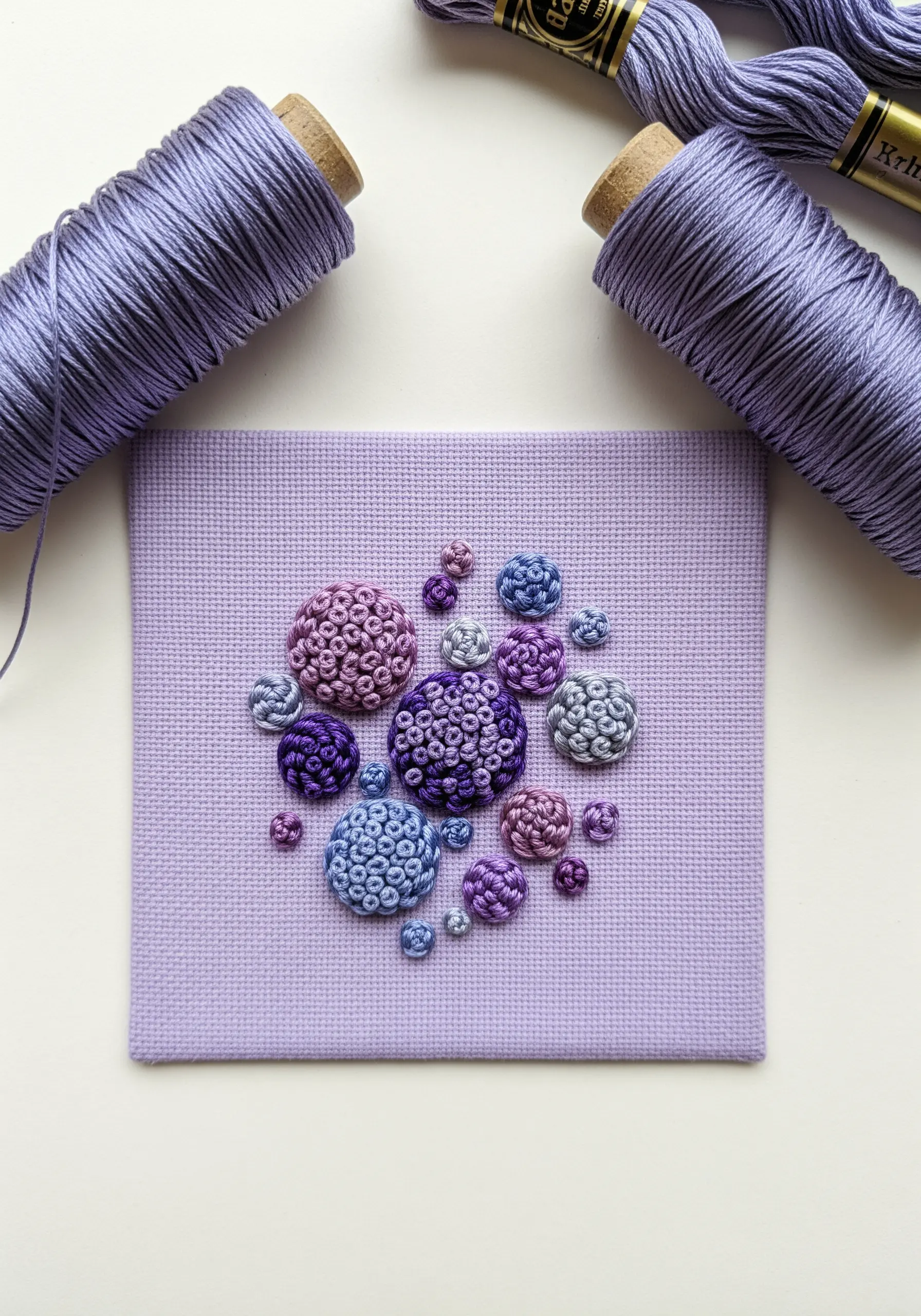 Clusters of French knots in varying shades of purple and blue on lavender fabric.