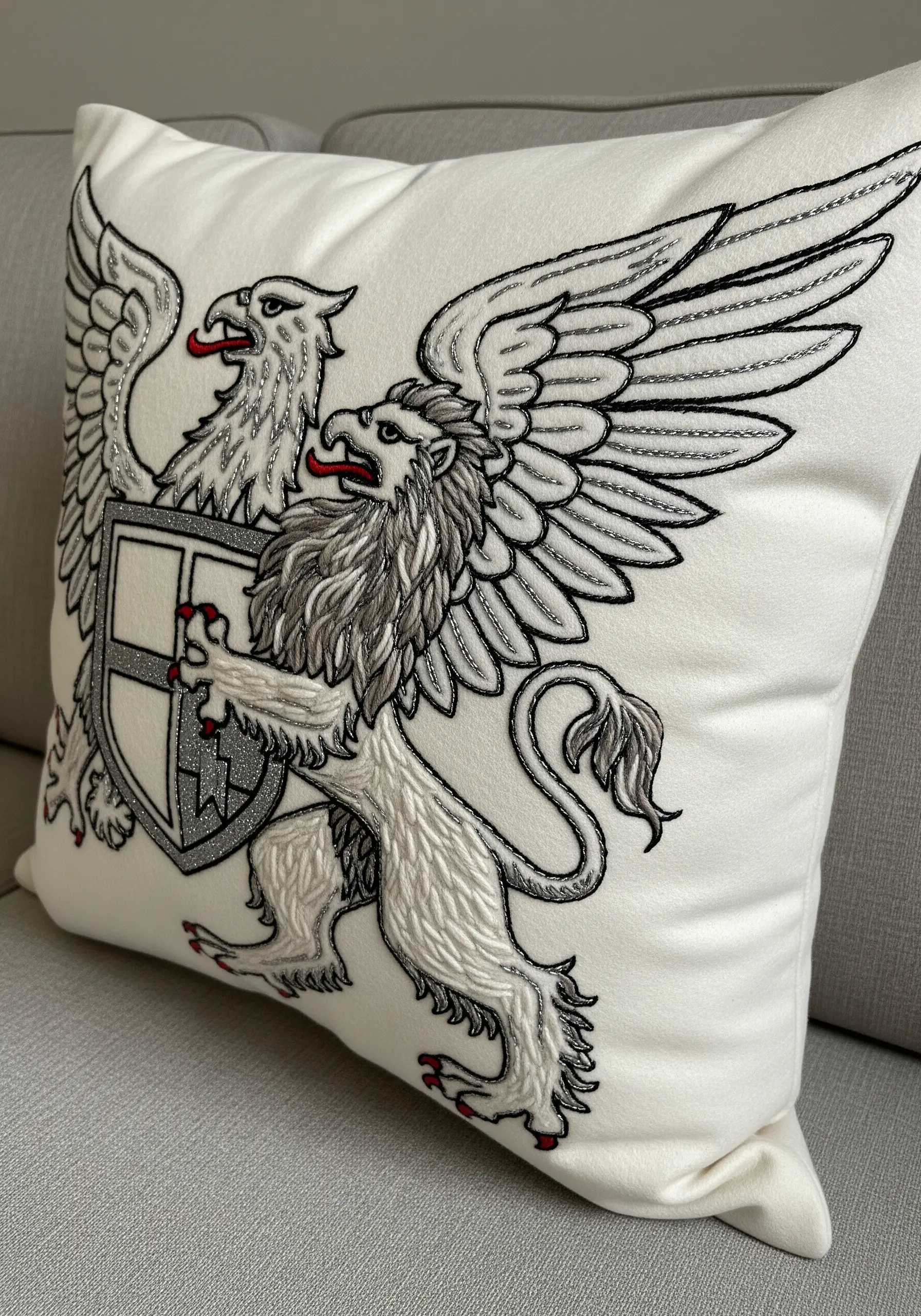 A monochrome embroidered griffin with silver and red accents on a white cushion.