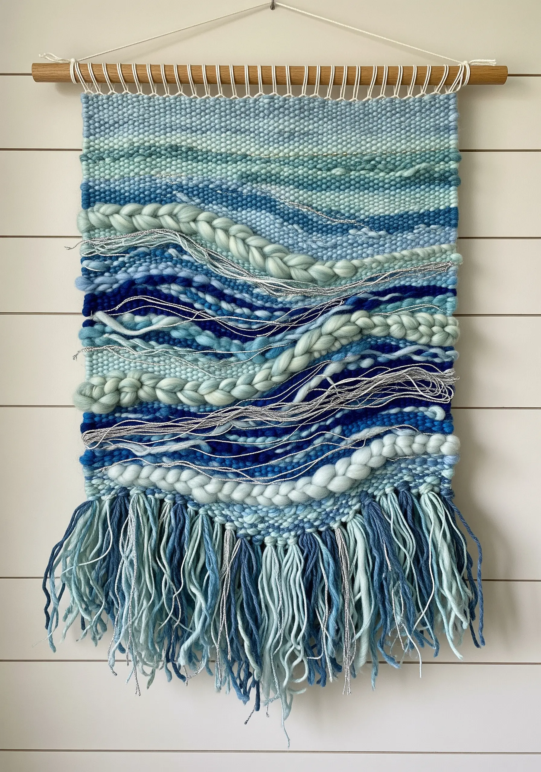 A woven wall hanging in shades of blue and seafoam green with varied textures and a long fringe.