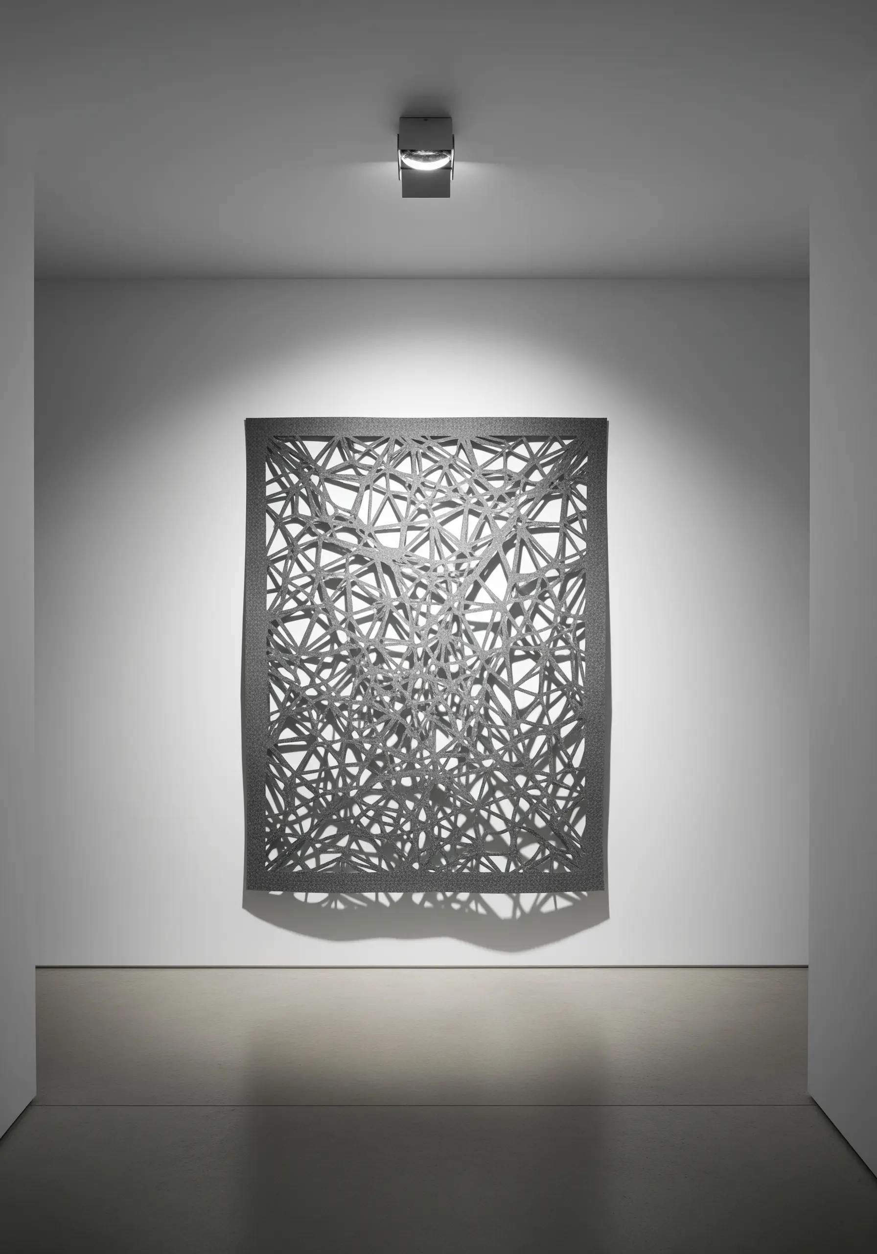 A square, web-like sculpture made of intersecting dark gray thread in a gallery.