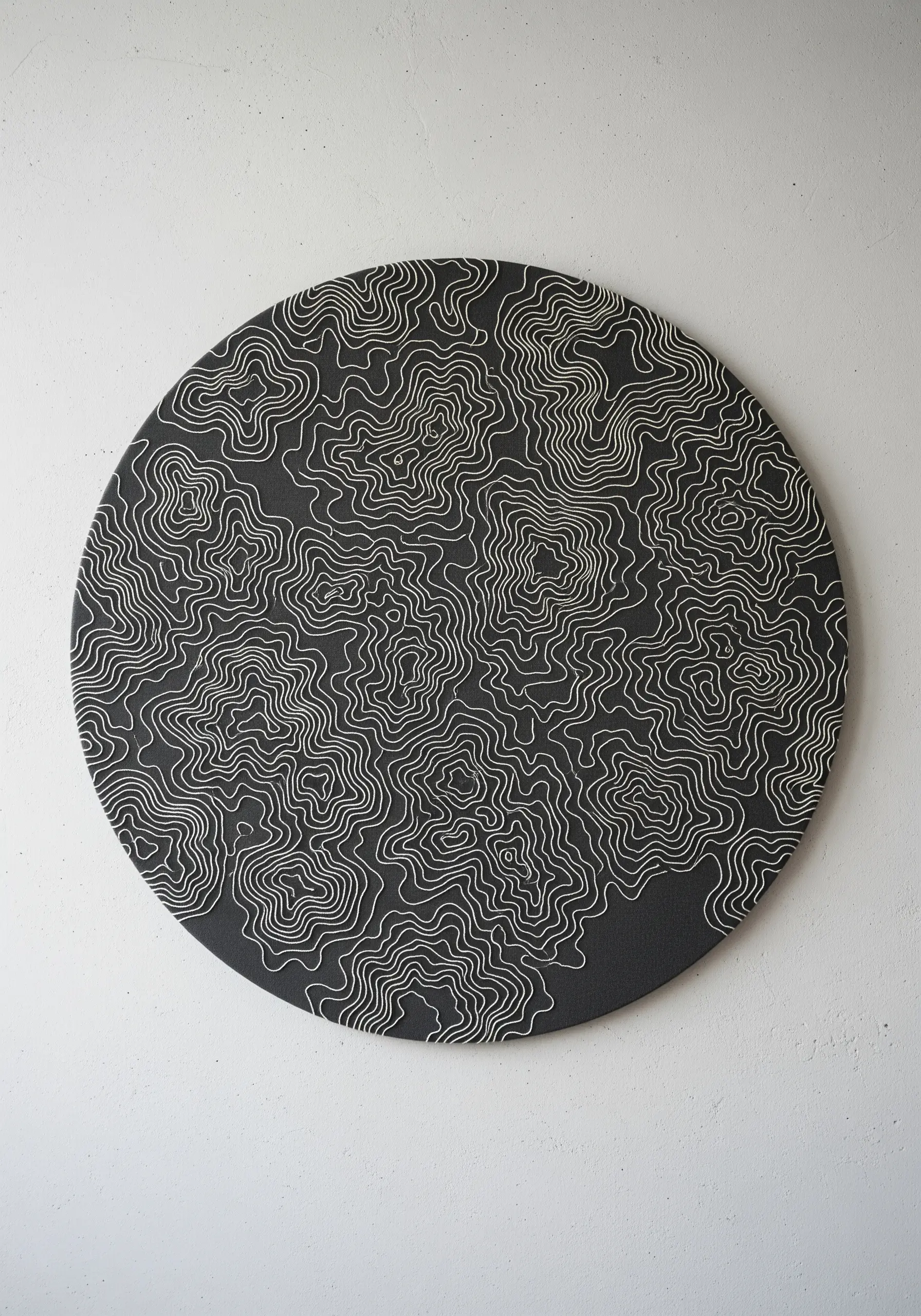 A dense, abstract pattern of topographic lines embroidered in white on a round black canvas.