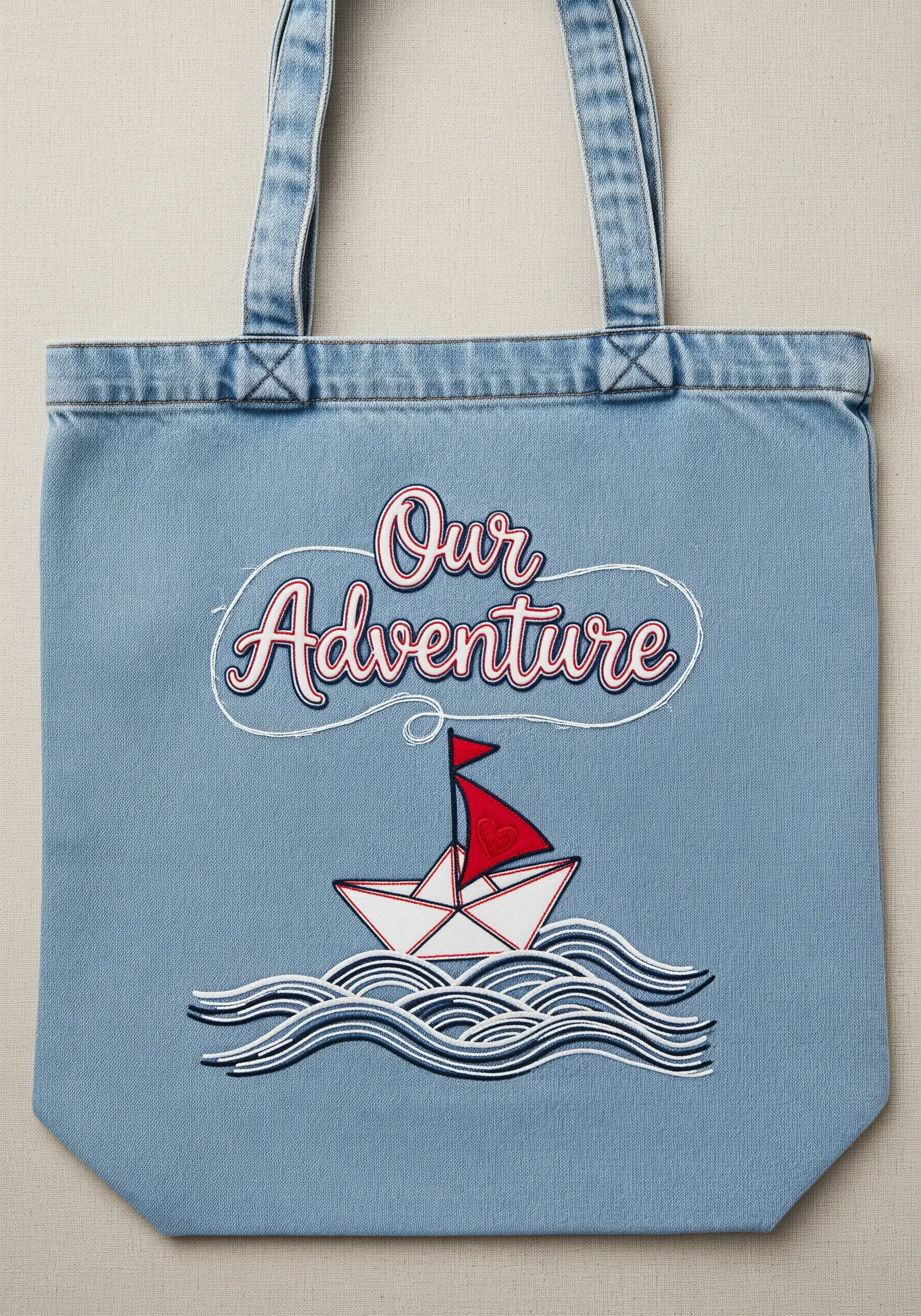 Embroidered 'Our Adventure' text above a paper boat on stylized waves, stitched on a denim tote.