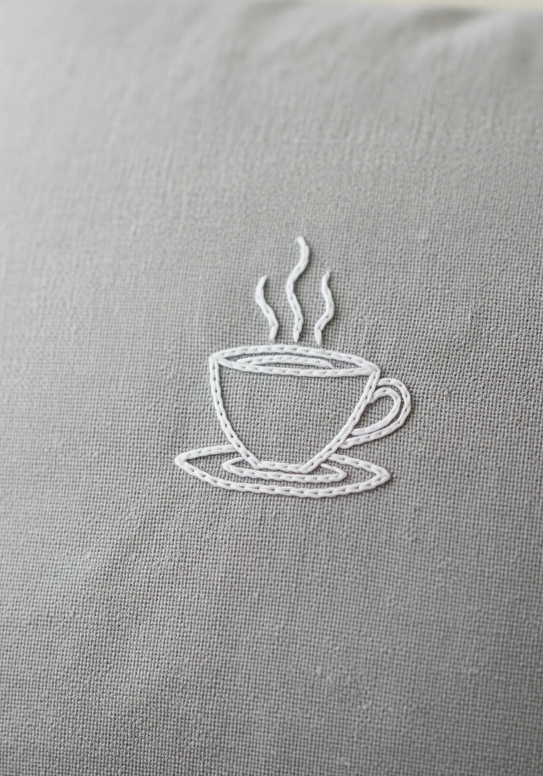 A simple outline of a steaming coffee cup embroidered in white thread on a gray linen pillowcase.