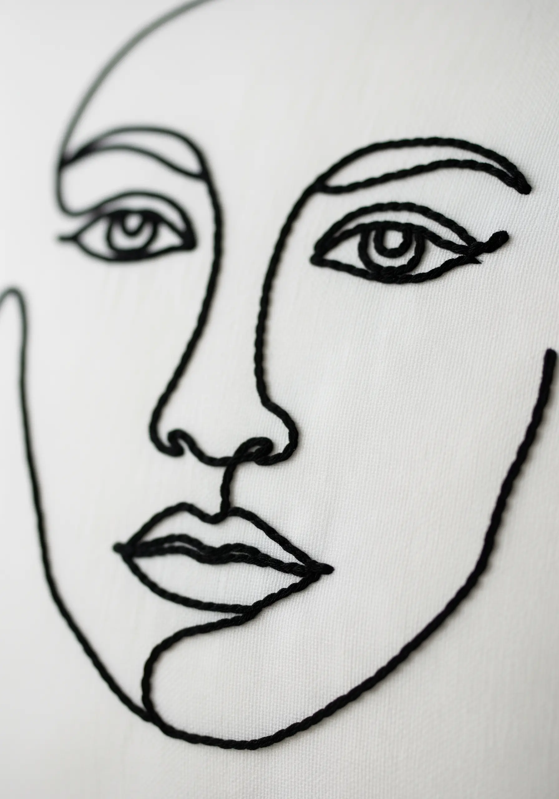 A close-up of a minimalist, abstract face embroidered in a single black line on a white pillowcase.