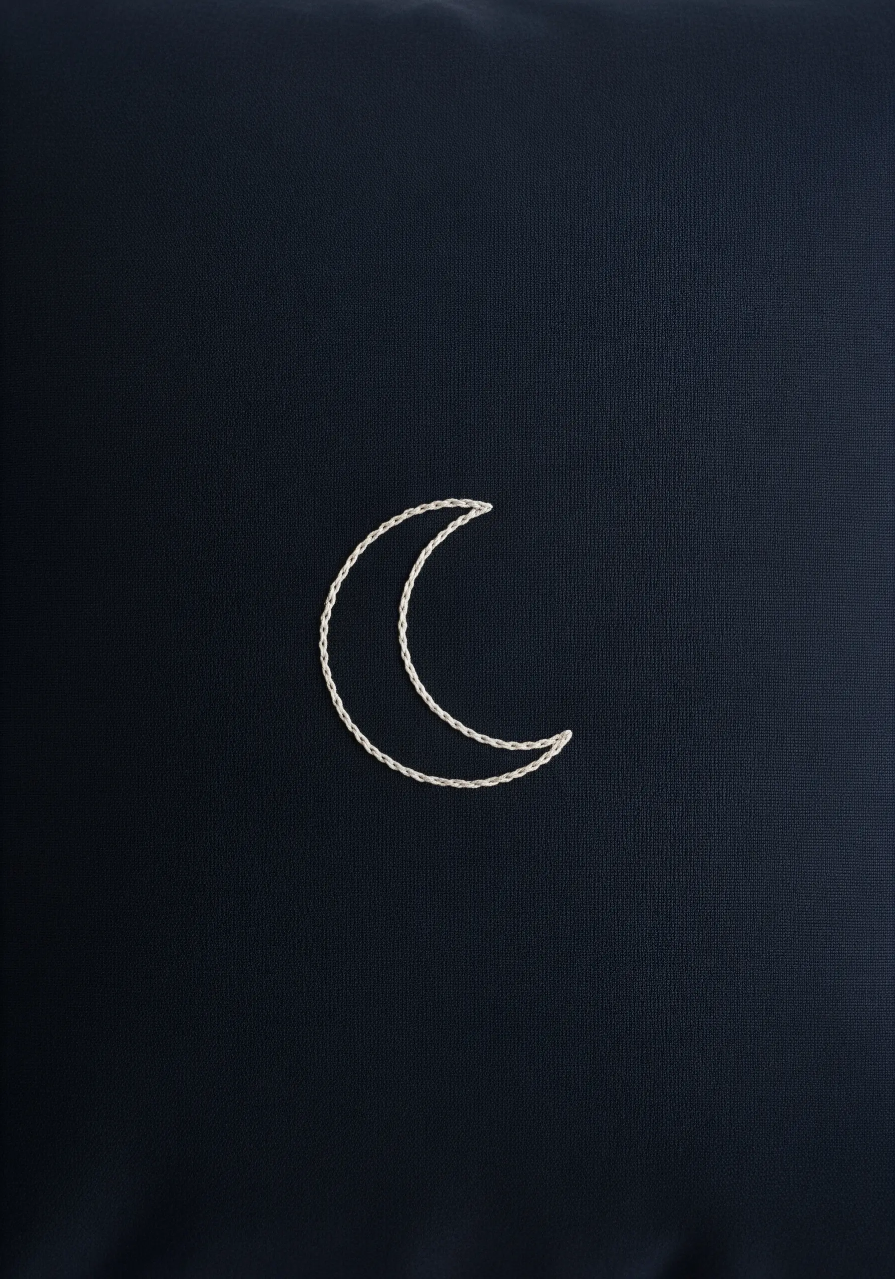 A simple crescent moon outlined in a white chain stitch on a dark navy blue pillowcase.