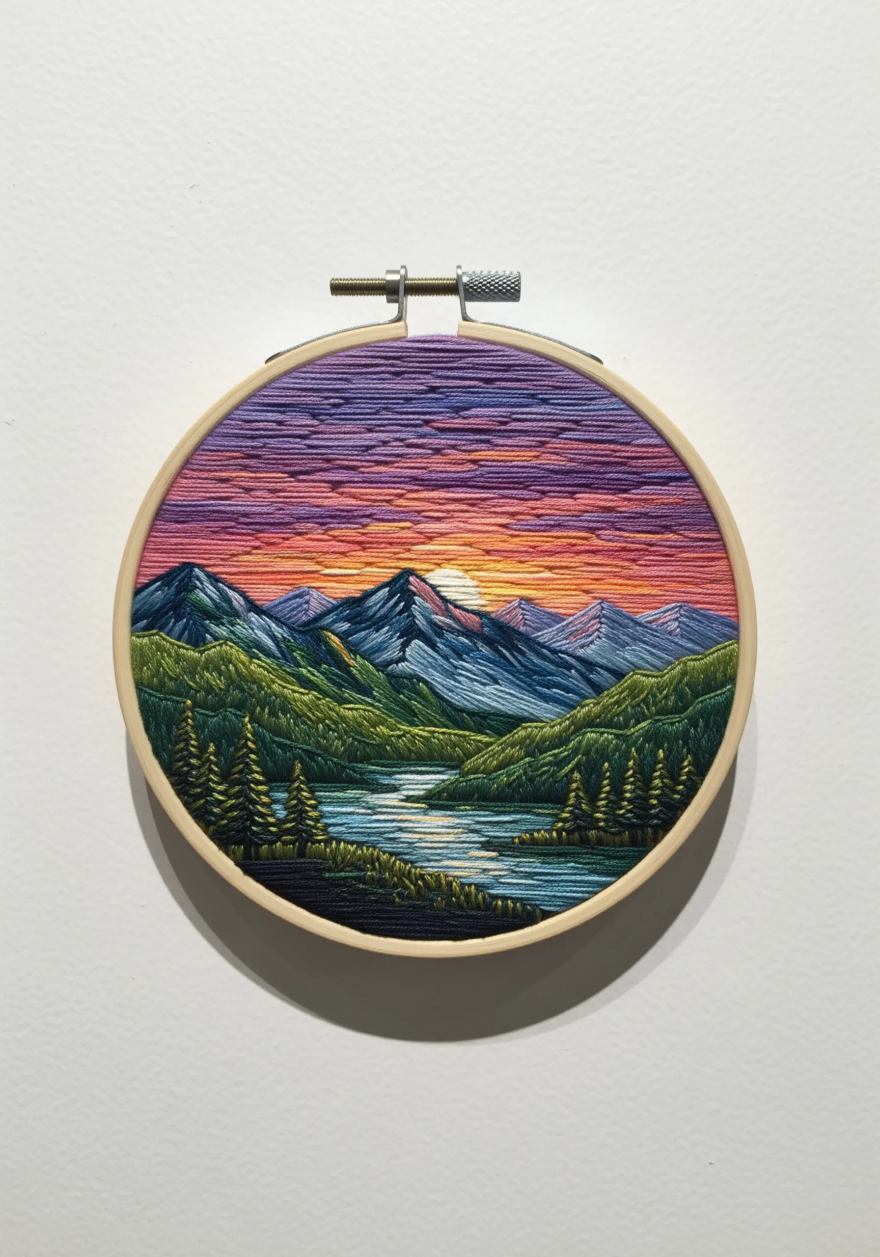 A detailed landscape embroidery in a hoop, depicting a mountain sunset over a river.