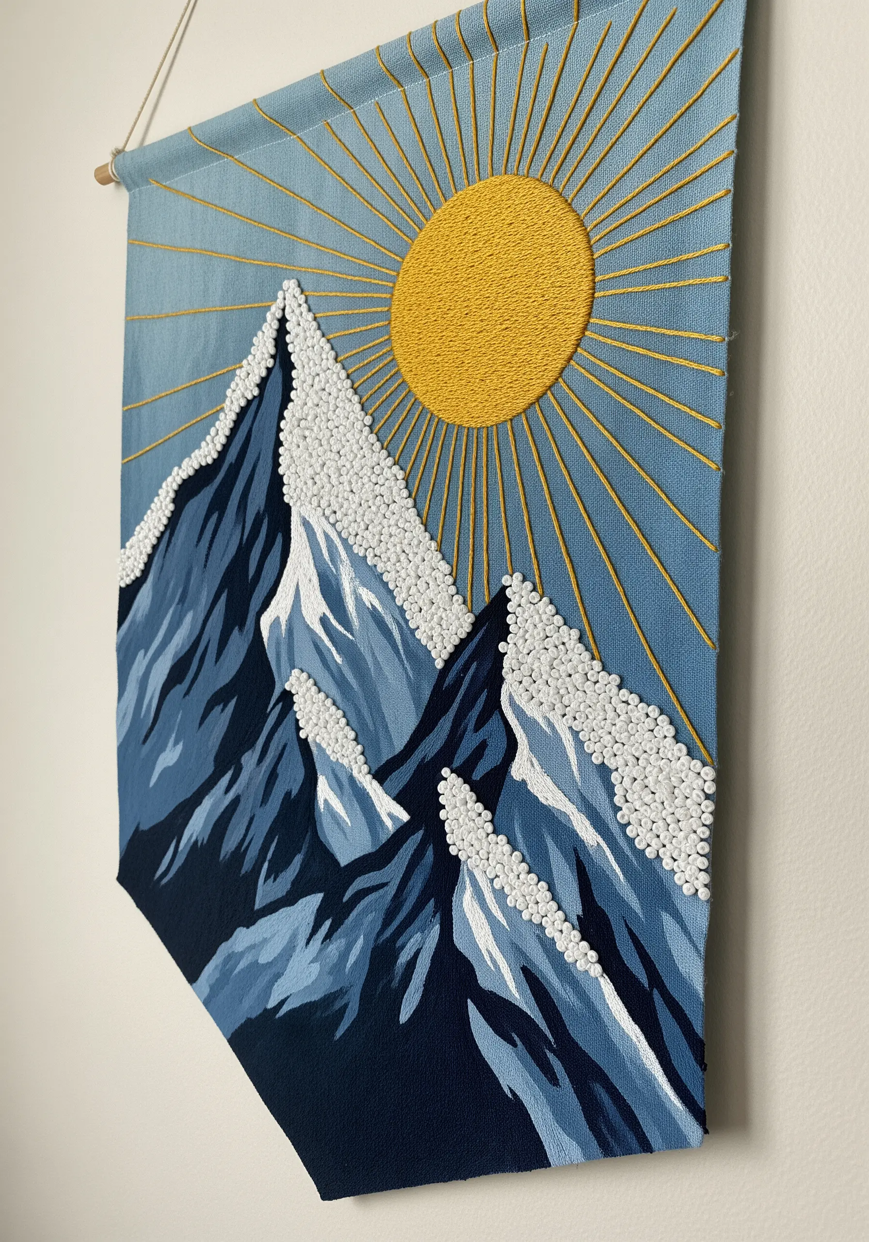 A wall hanging of mountains with a painted background and embroidered sun and snowy peaks.