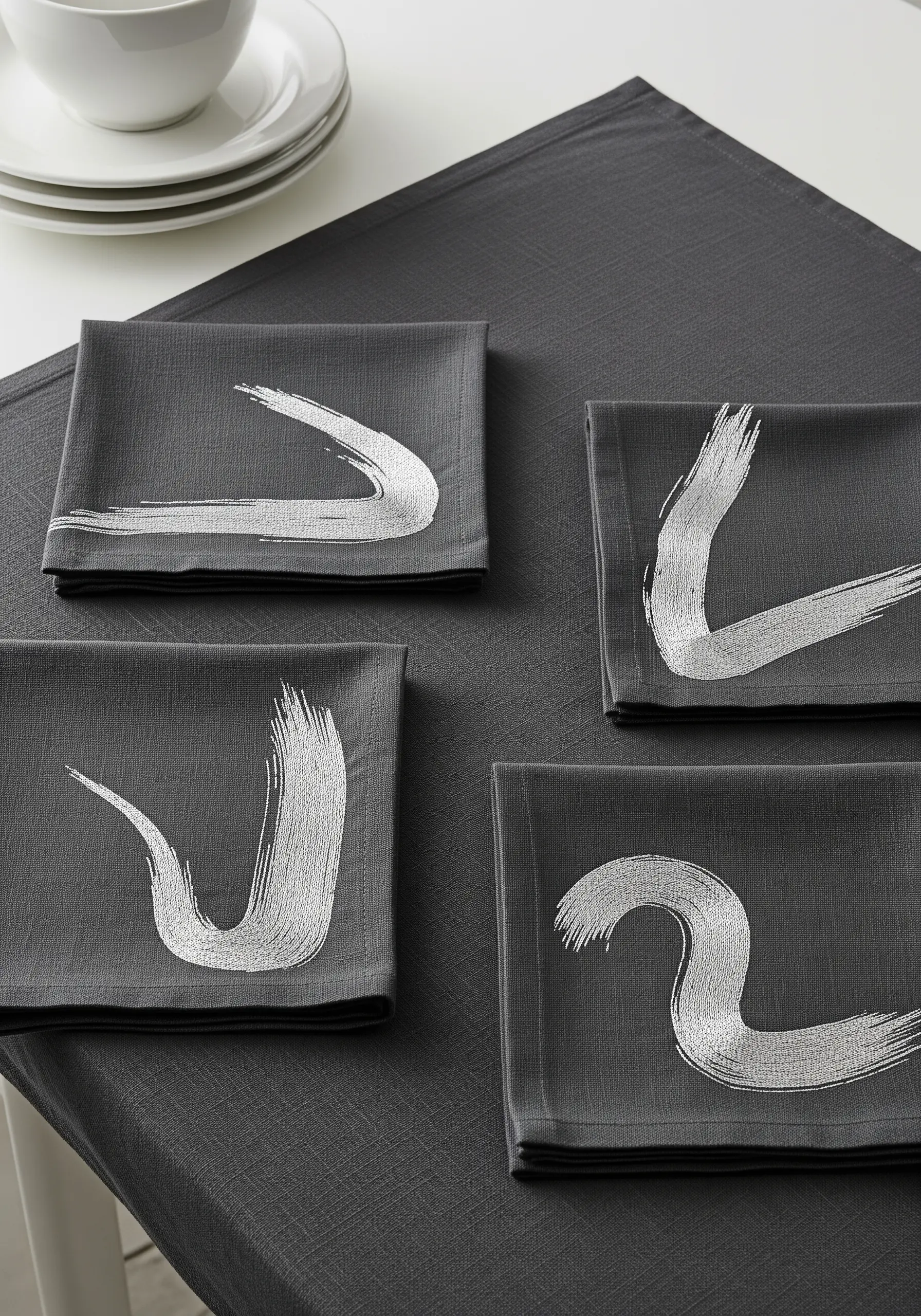 Four dark grey napkins, each embroidered with a single, expressive silver metallic brushstroke.