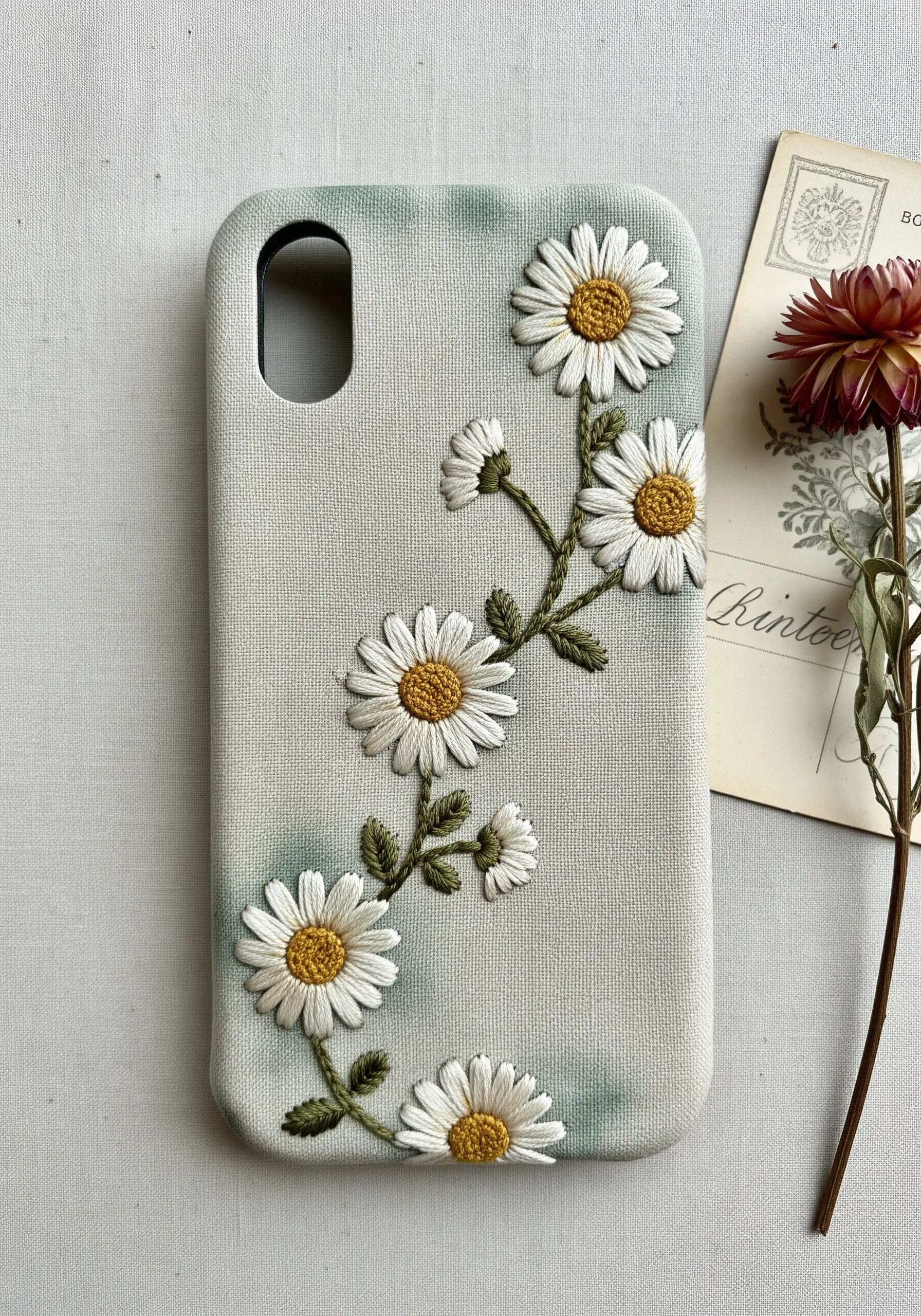 Realistic daisies embroidered on a phone case with blended, painterly petals and variegated stems.