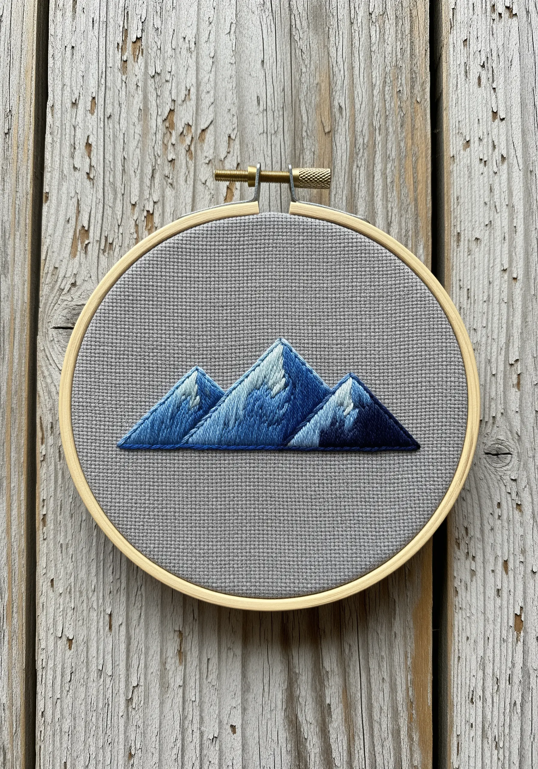 A mountain range embroidered with blue and white thread painting on grey Aida cloth.