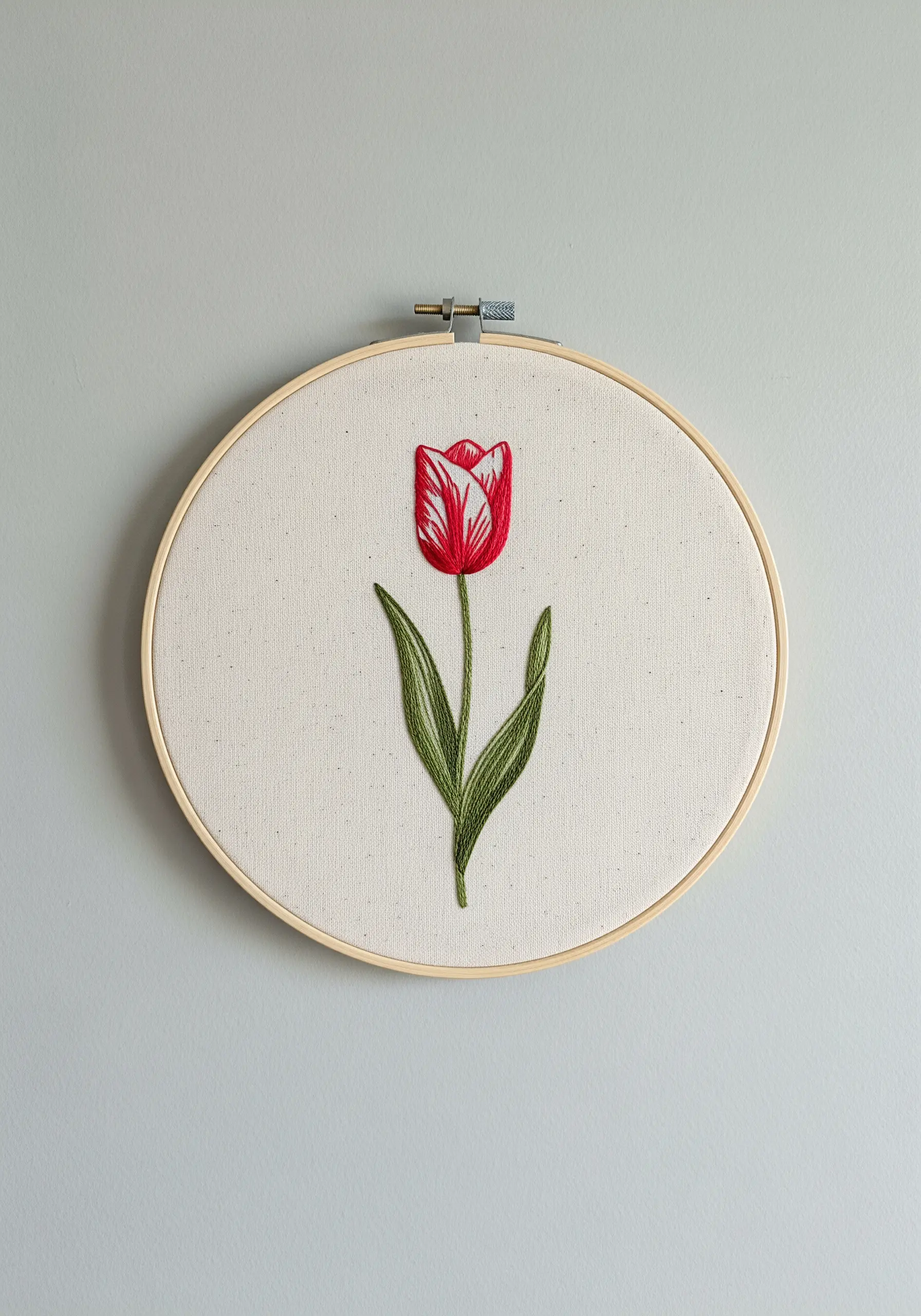 A realistically shaded red and white tulip with green leaves, embroidered in a hoop.